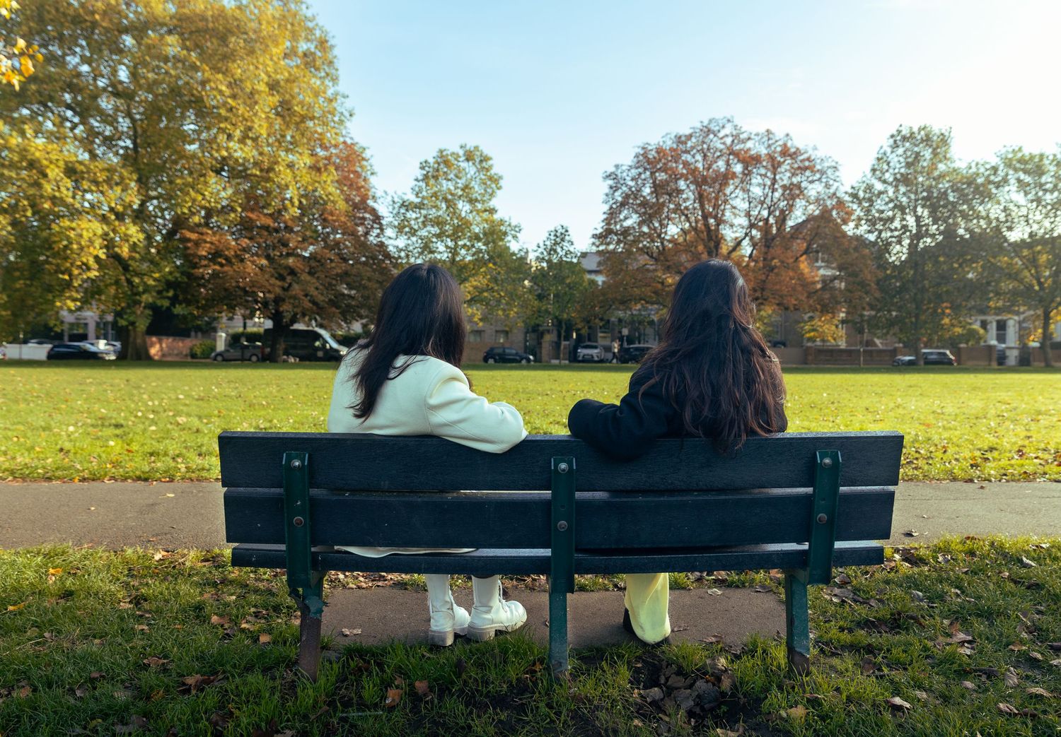 Two women with dark hair sitting side by side on a park bench surrounded by autumn foliage.
