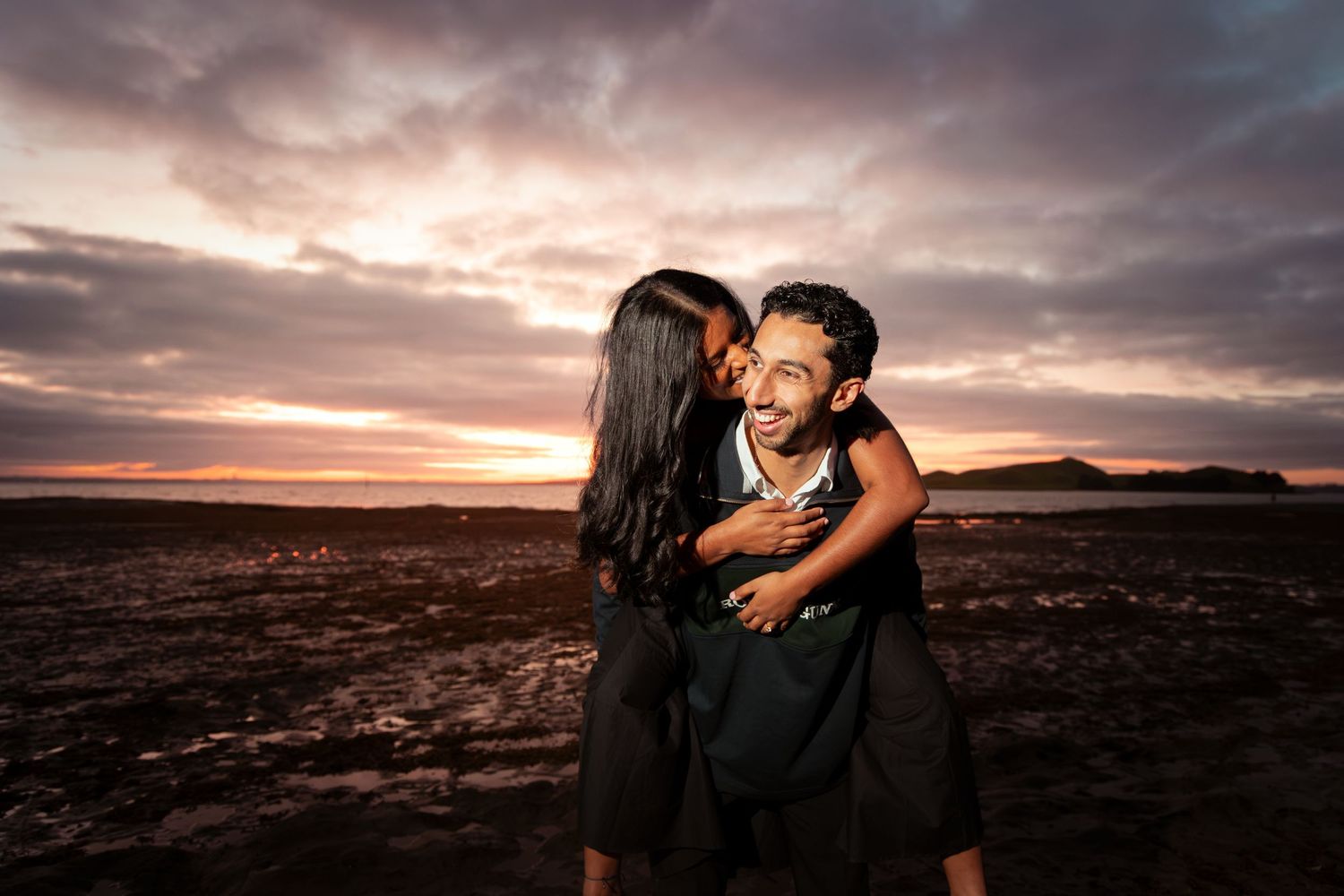 Couple embracing romantically at sunset on a beach, woman kissing man's cheek with dramatic cloudy sky.