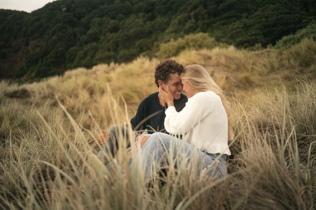 Romantic couple sitting together in golden grass field with lush green hills in the background.