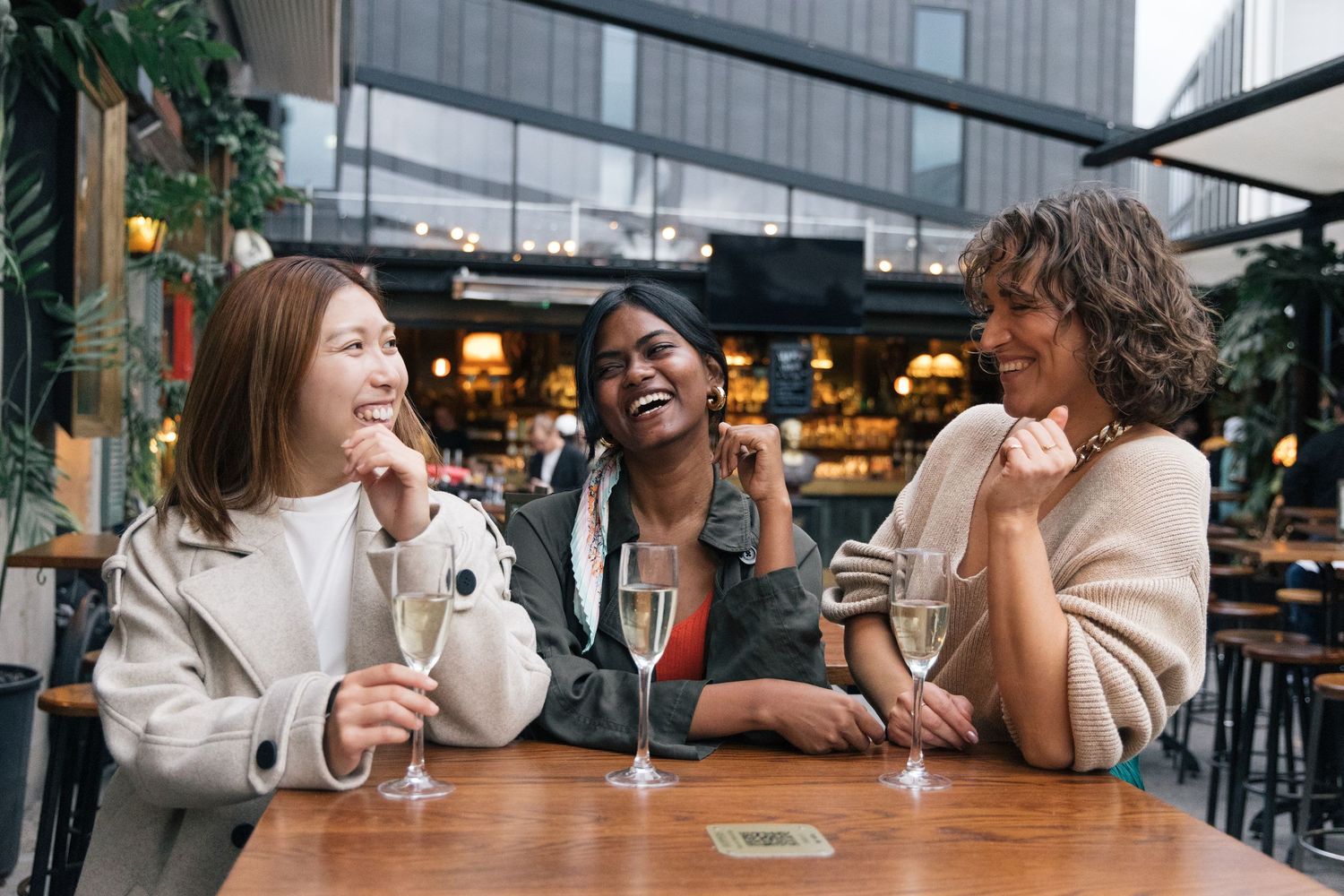 Three diverse women laughing and enjoying champagne drinks at an outdoor bar table.
