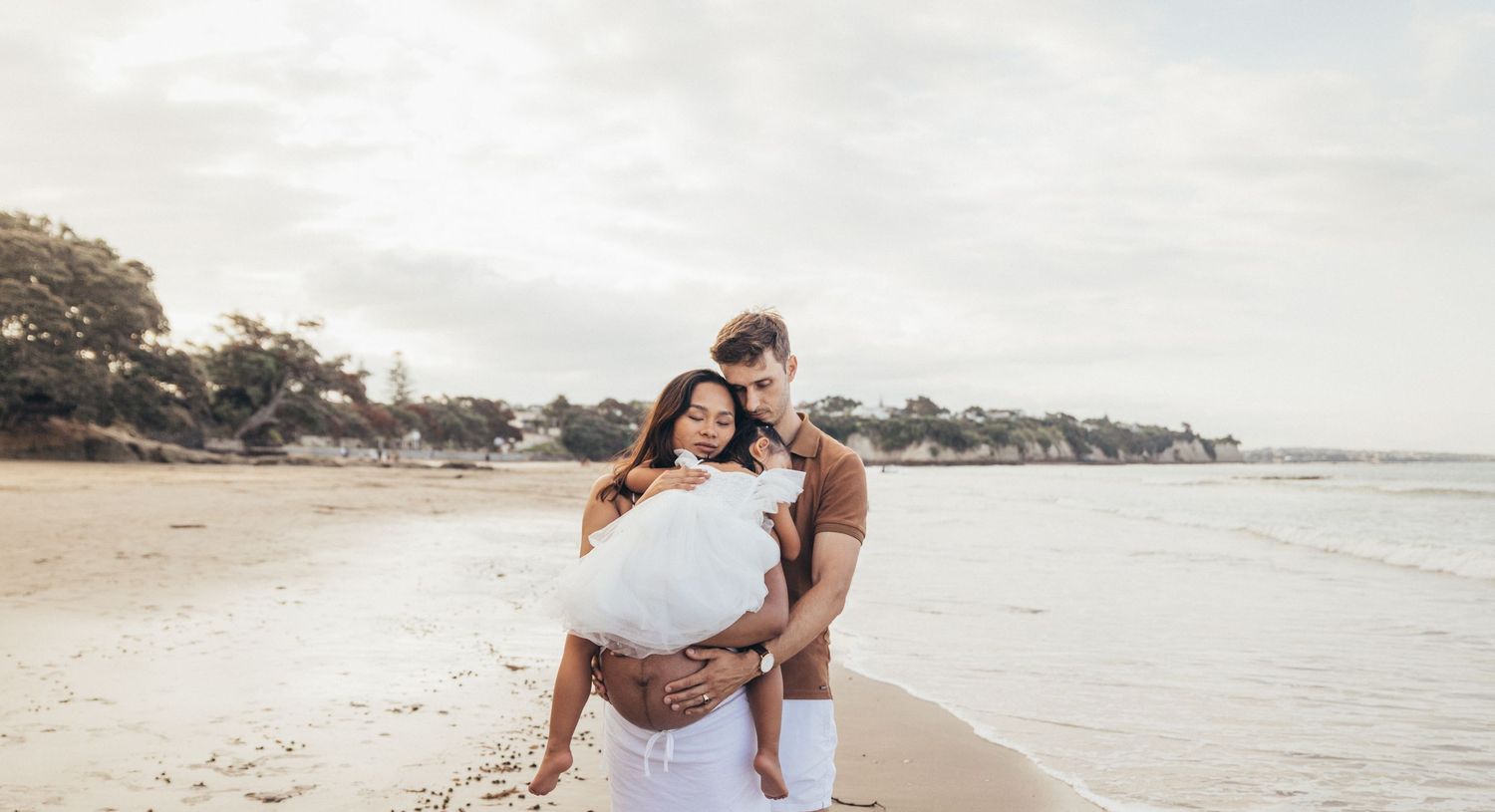 Couple embracing on a sandy beach, woman in white dress, romantic outdoor maternity photo session.