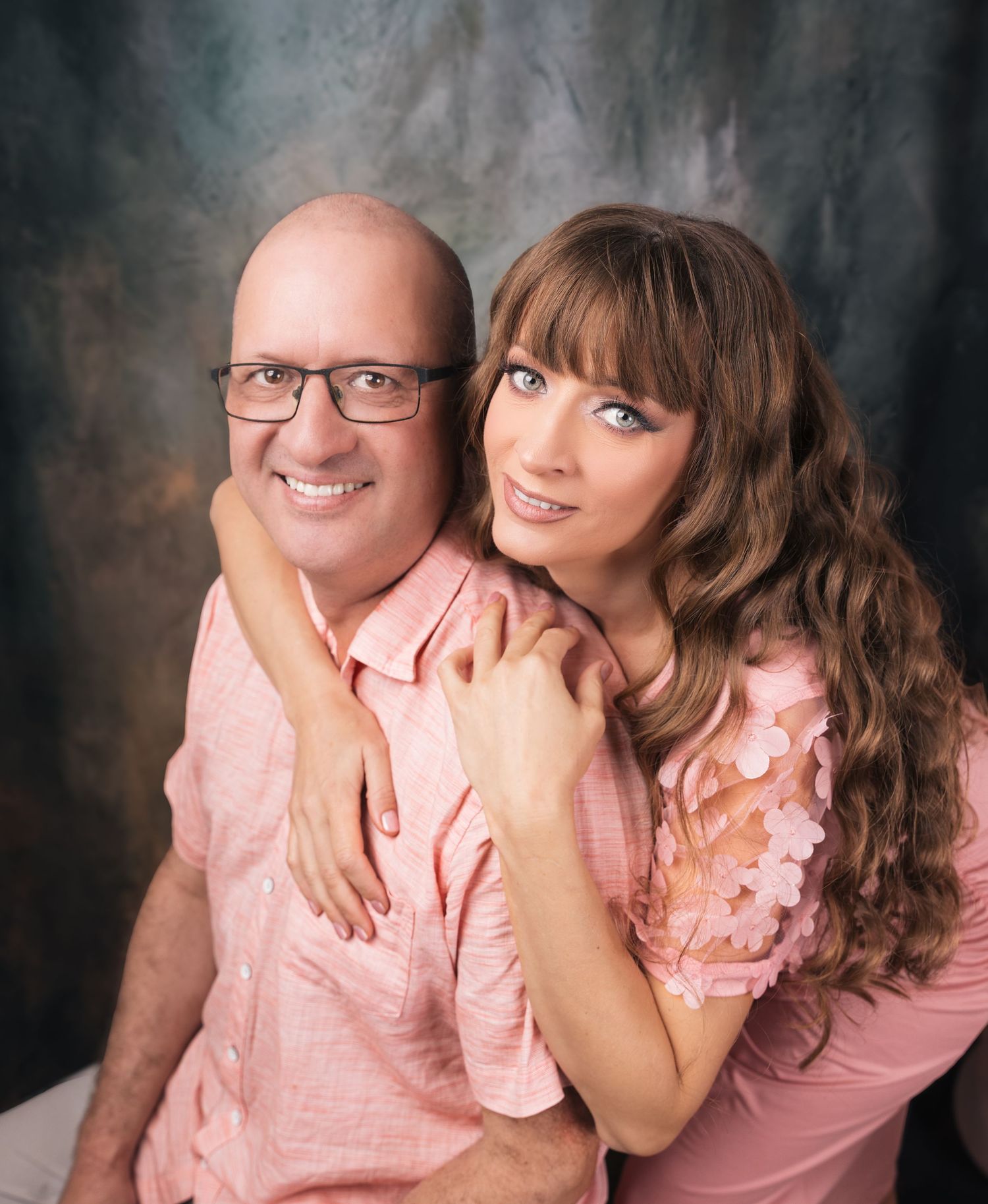 A smiling couple in matching pink outfits posing together against a mottled gray studio backdrop.