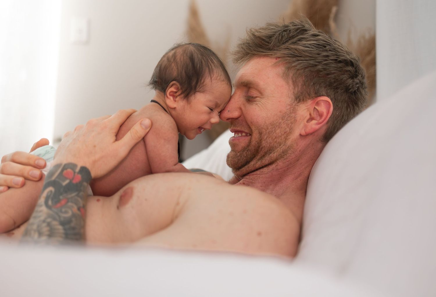 A tattooed man lying in bed smiling while holding a newborn baby close to his face in a tender moment.