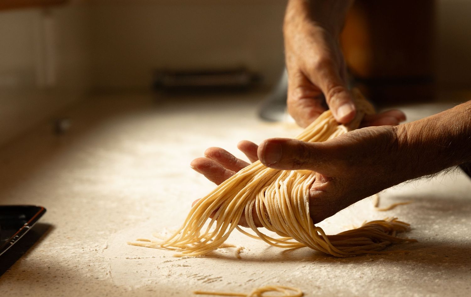 Hands holding freshly made spaghetti nests on a flour-dusted surface in warm light.