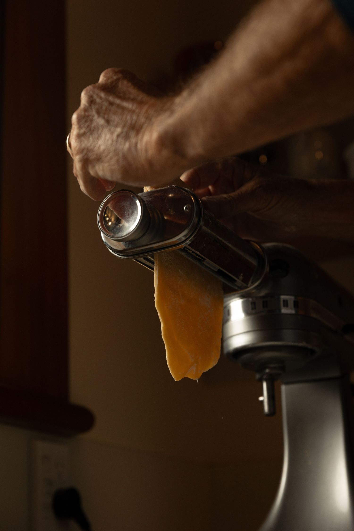 Hands feeding pasta dough through a KitchenAid pasta roller attachment in a dark kitchen.