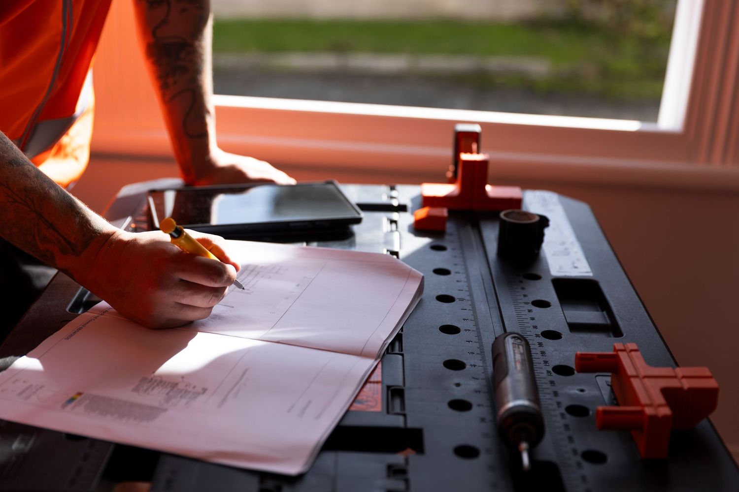 Construction worker in orange vest writing on blueprints at a workbench with tools.