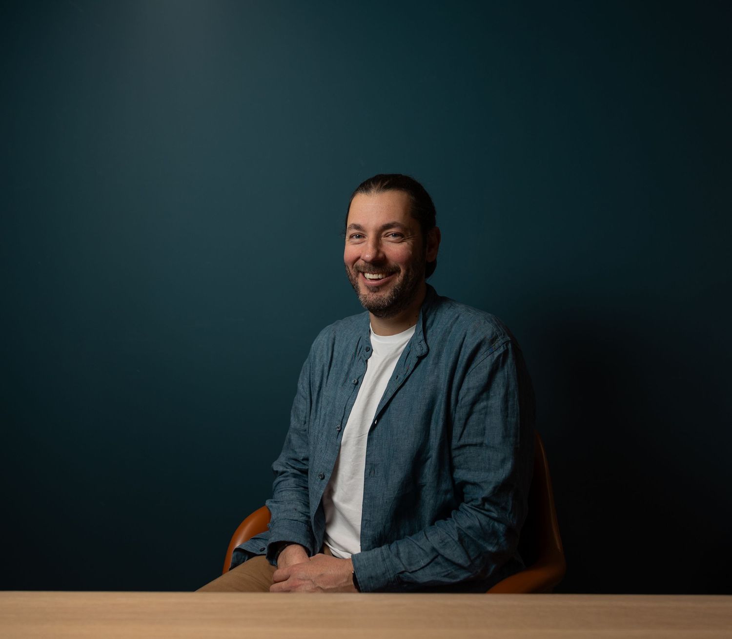 Cheerful man in a blue denim shirt sitting at a wooden table against a teal background.