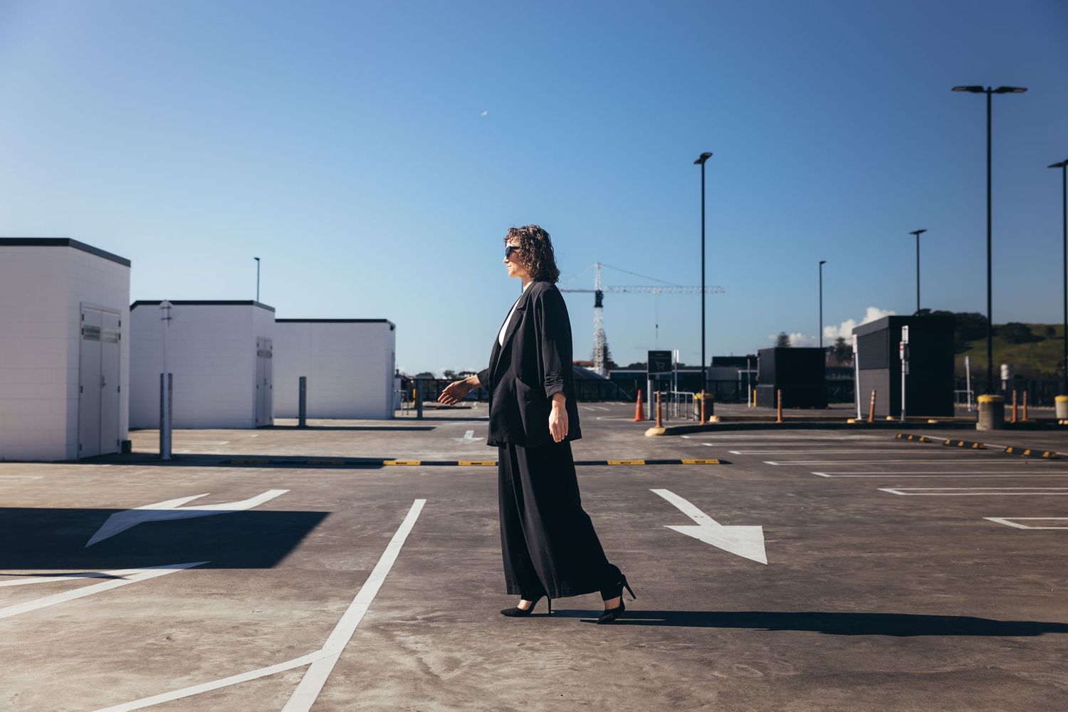 Stylish woman in long black dress walking through open parking lot under clear blue sky.