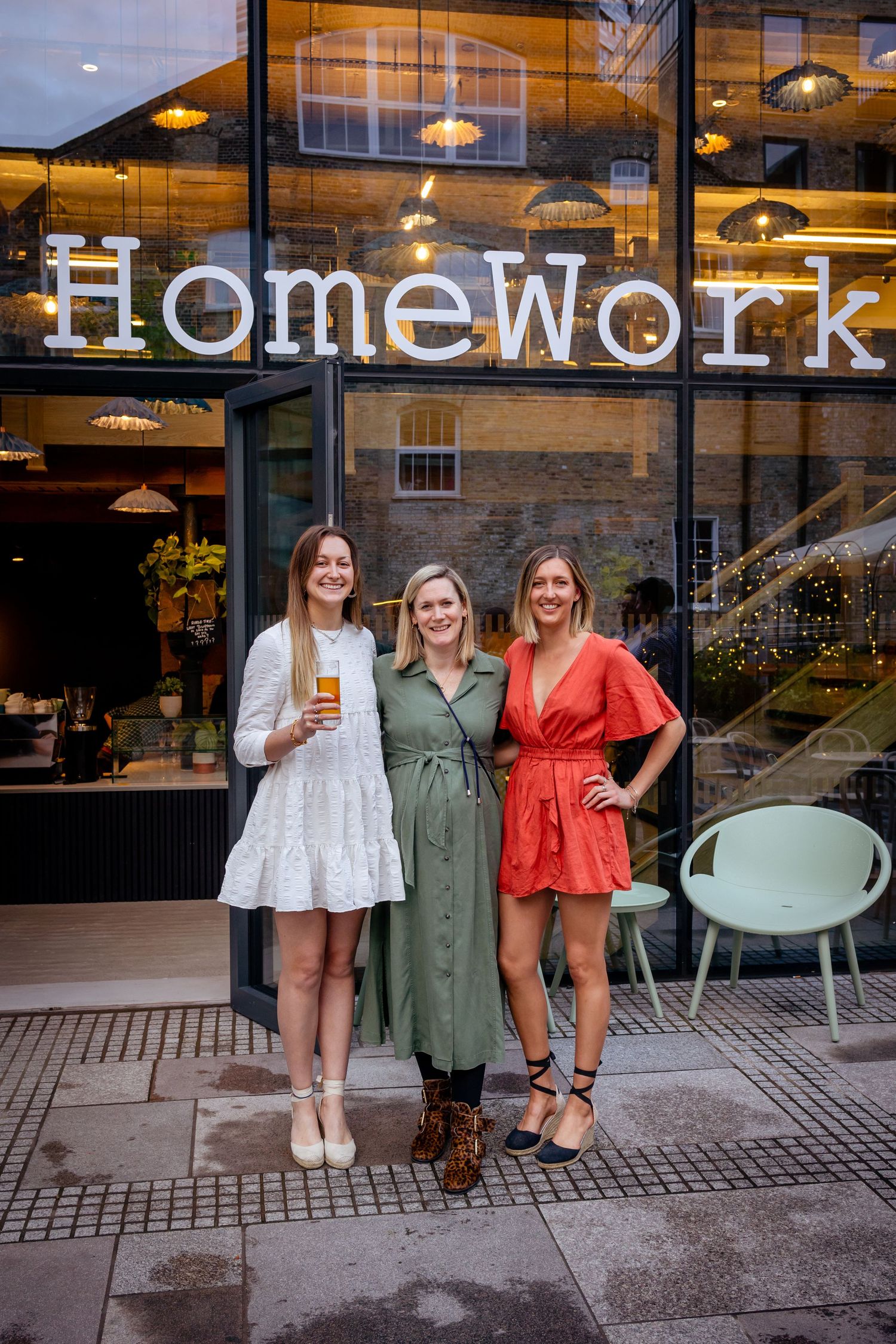 Three women smile together outside HomeWork, dressed in white mini, green midi, and red wrap dresses.