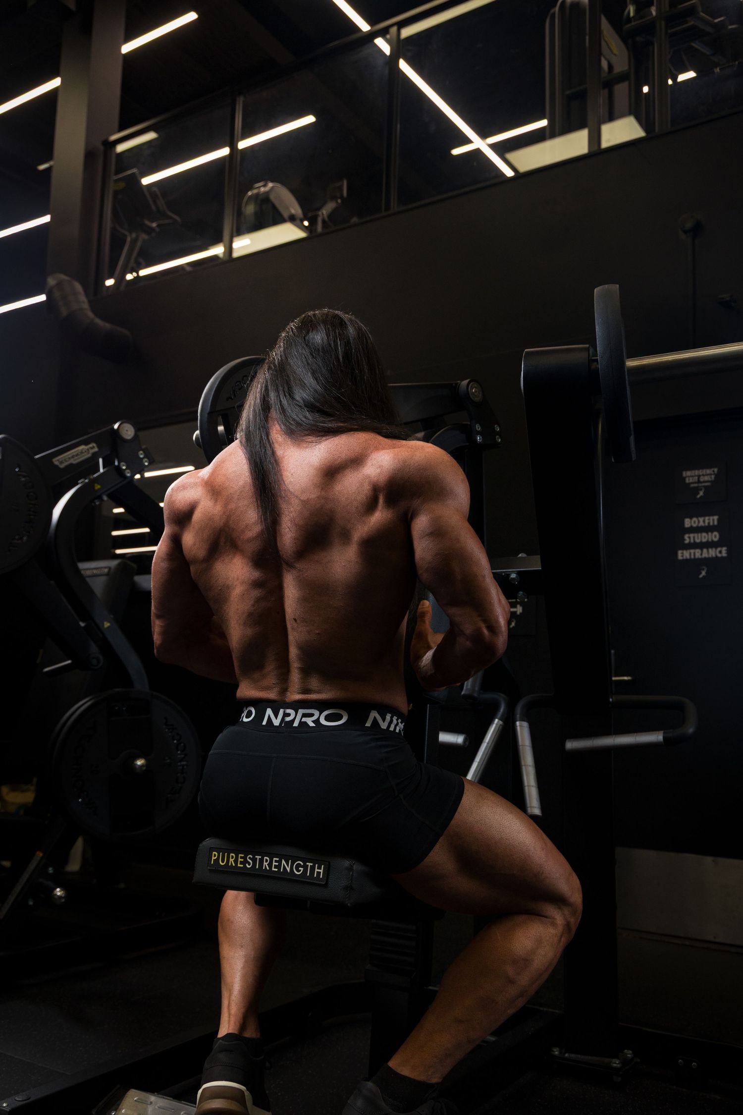 Muscular bodybuilder training back muscles on gym equipment, wearing Nike Pro shorts, rear view in dark gym.