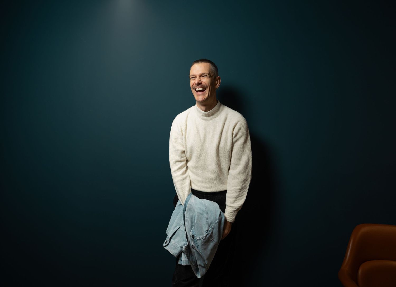 Man in a white turtleneck sweater laughing while standing against a dark teal wall.