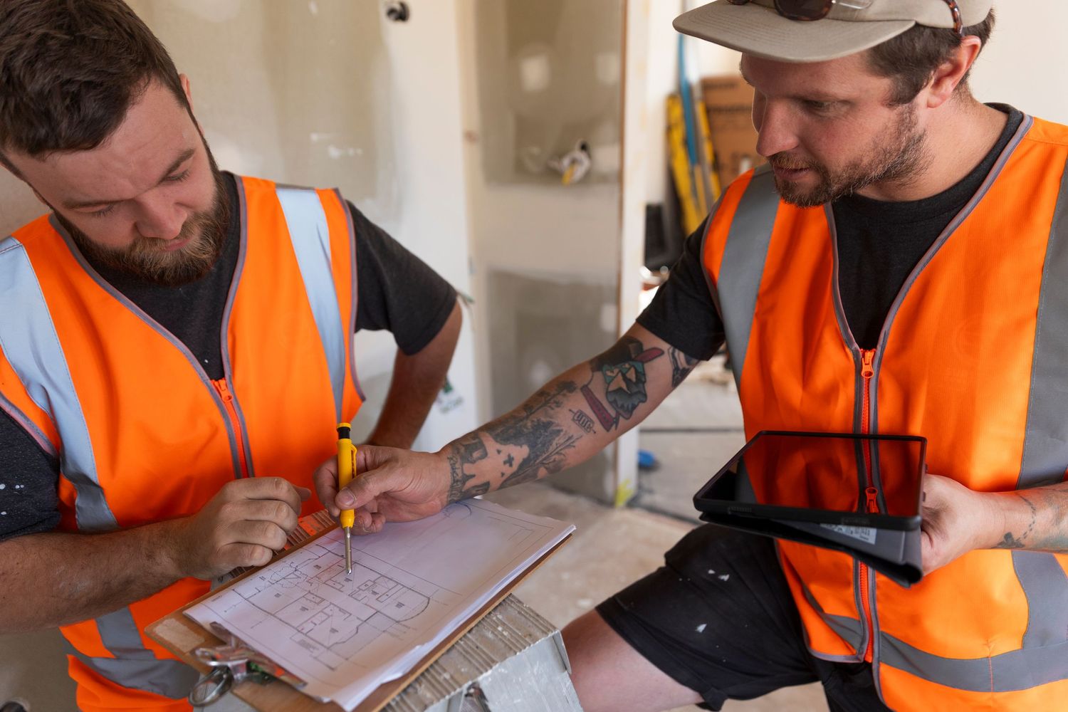 Two tattooed builders in orange safety vests reviewing floor plans with a tablet on site.