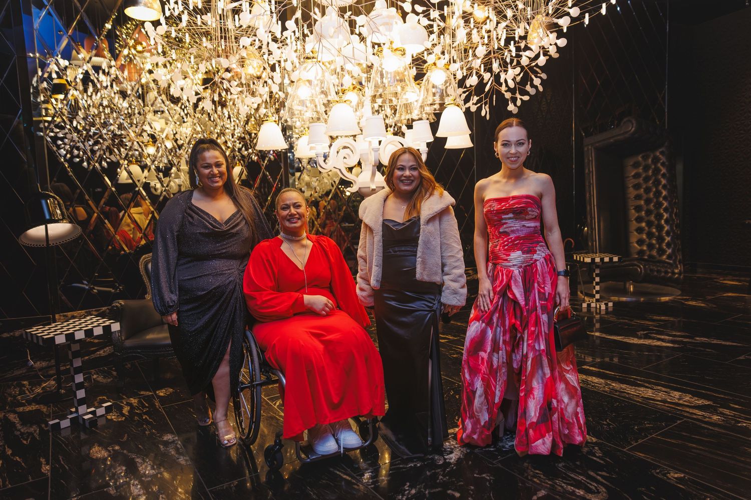 Four women posing together beneath decorative lights, including one seated in a wheelchair, at an elegant event.