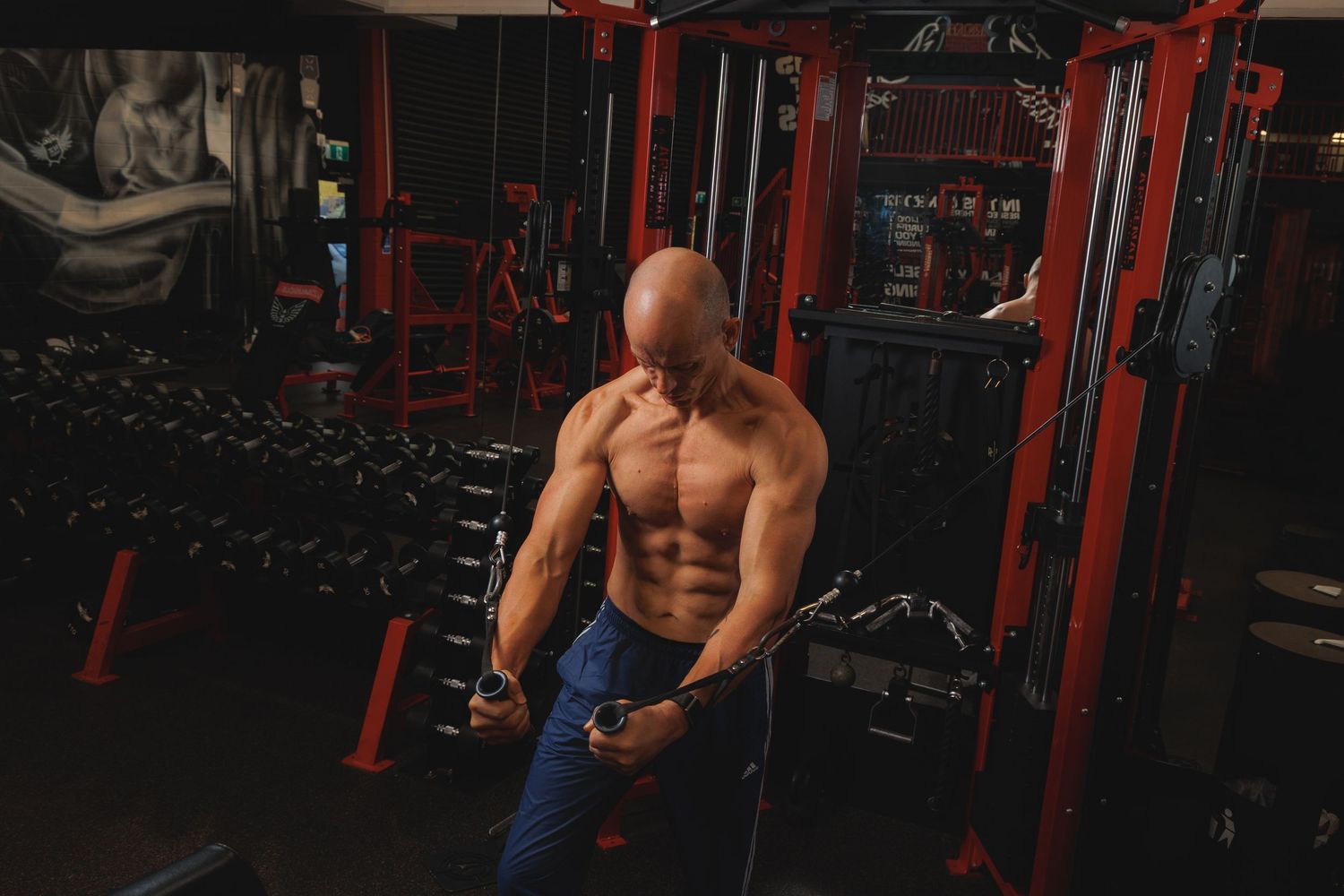 Bald muscular man doing cable exercises shirtless in a dark gym with red equipment.
