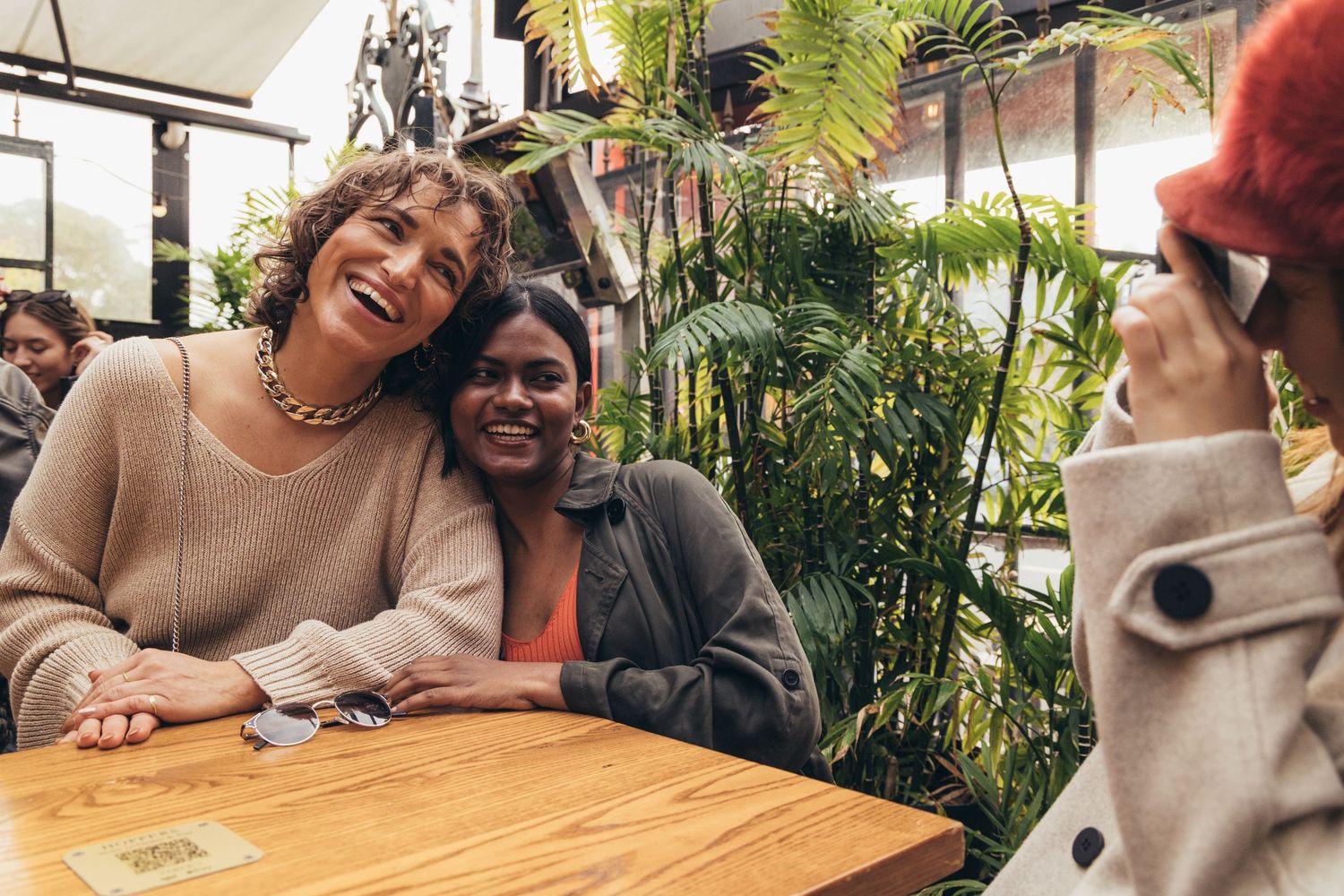 Two smiling women posing for a photo at a wooden table in a plant-filled cafe.