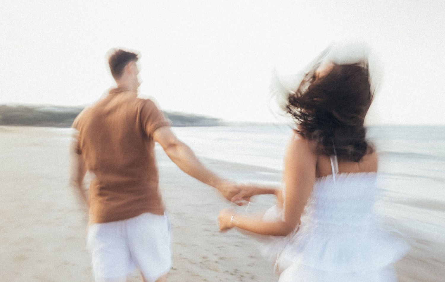 Couple holding hands running on a sandy beach, motion blur capturing romantic playful moment with ocean in background.