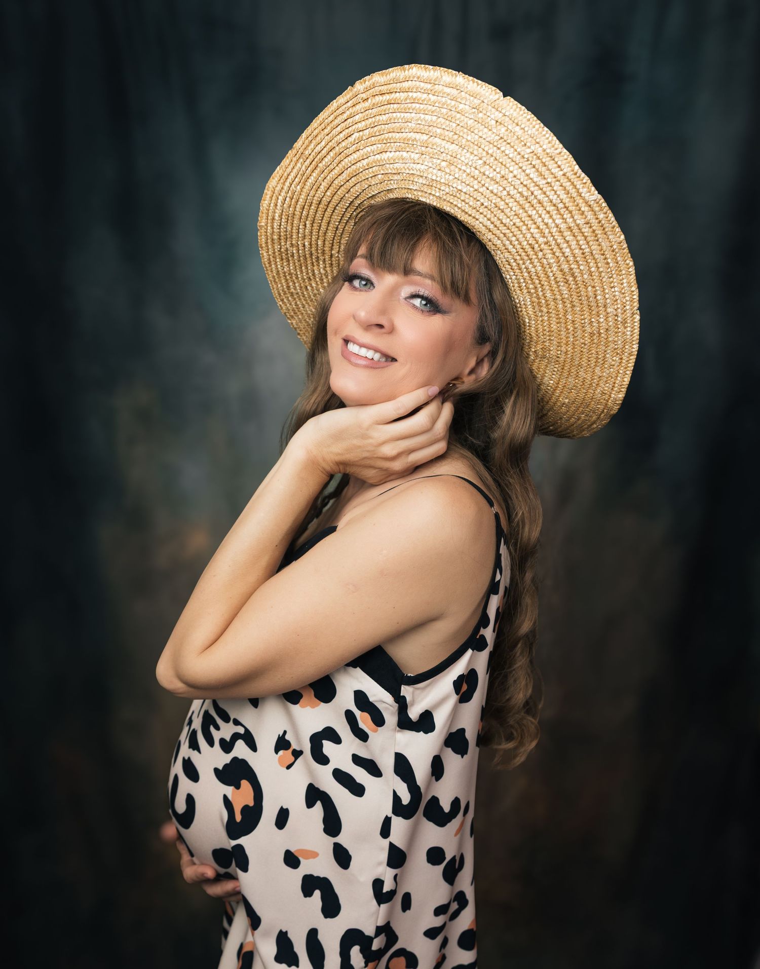 Smiling pregnant woman in straw hat and leopard print dress posing for maternity portrait photo.