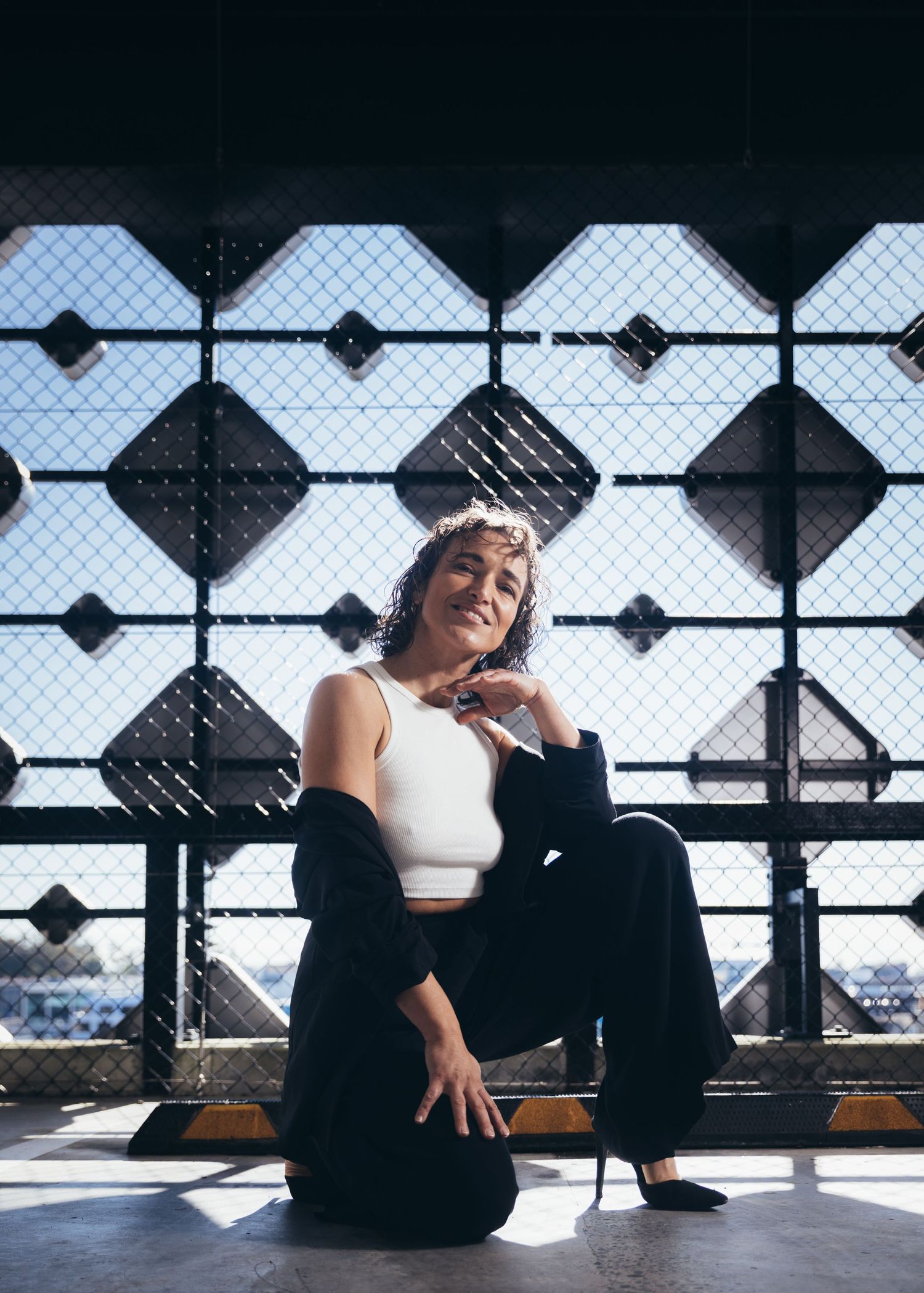Woman in white crop top and black pants sitting relaxed against geometric metal fence in sunlight.