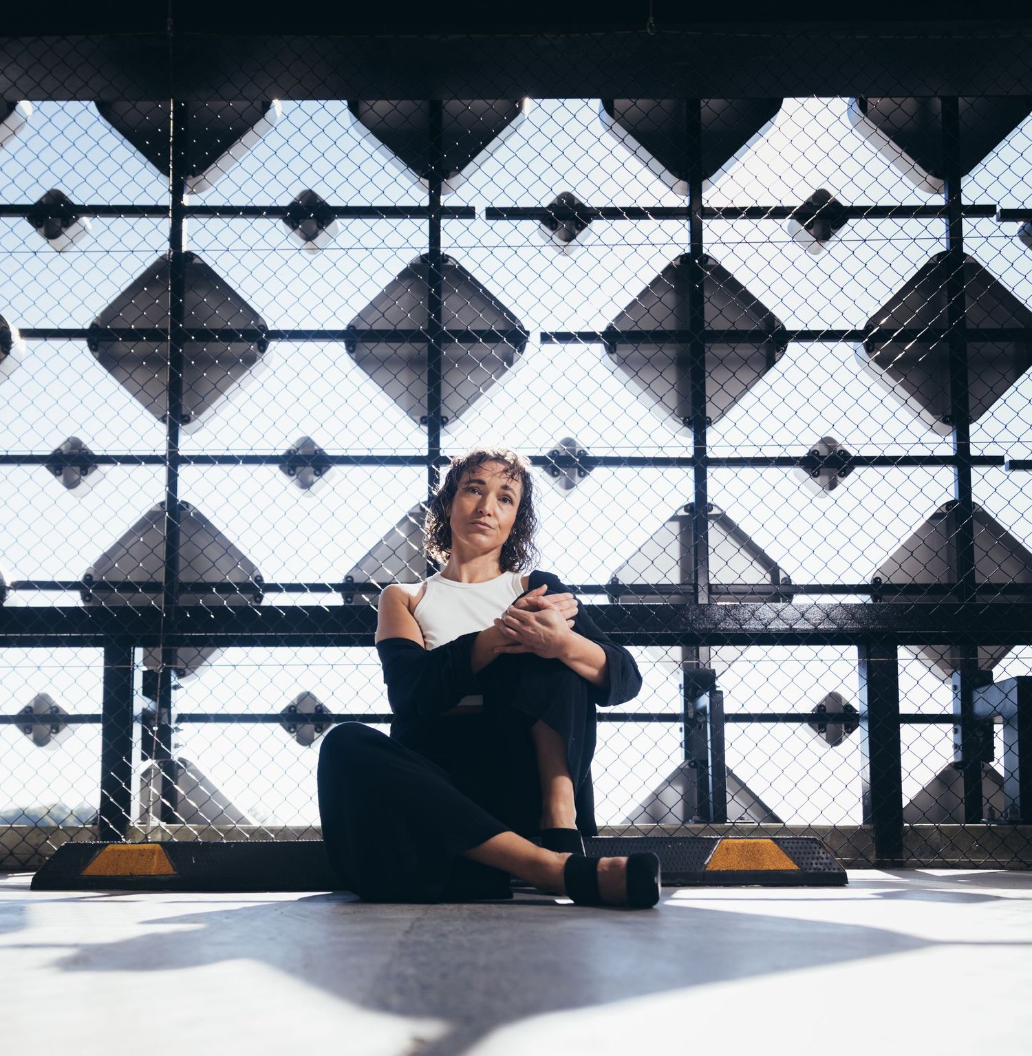 Woman in black outfit sitting cross-legged reading a book against geometric metal fence in sunlight.