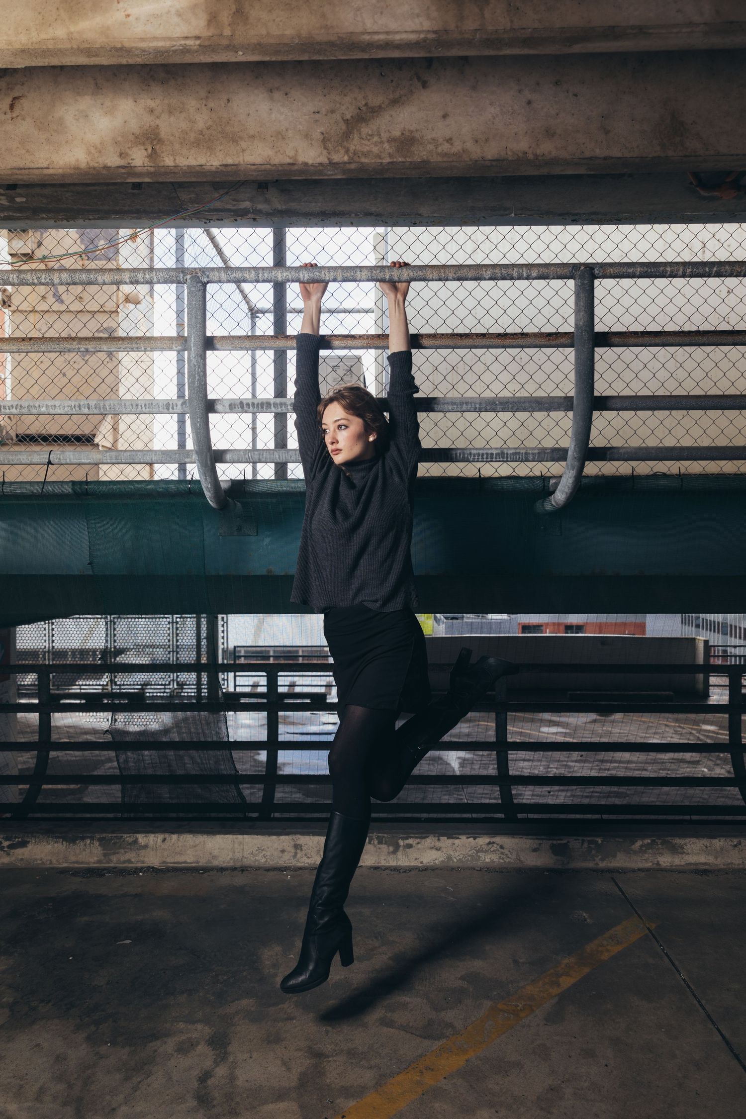Stylish woman in all-black ensemble hanging from metal fence in industrial parking garage setting.