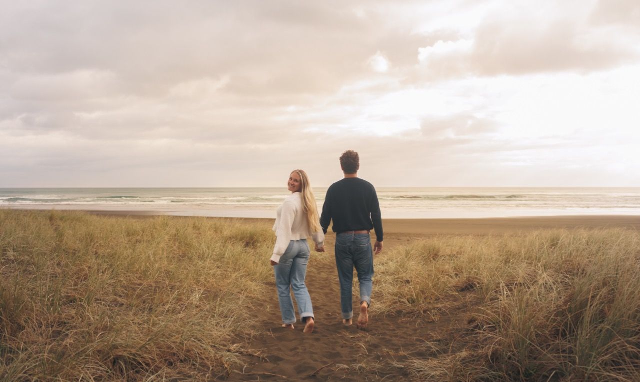 Couple holding hands walking toward ocean beach through golden grass under cloudy sky at sunset.