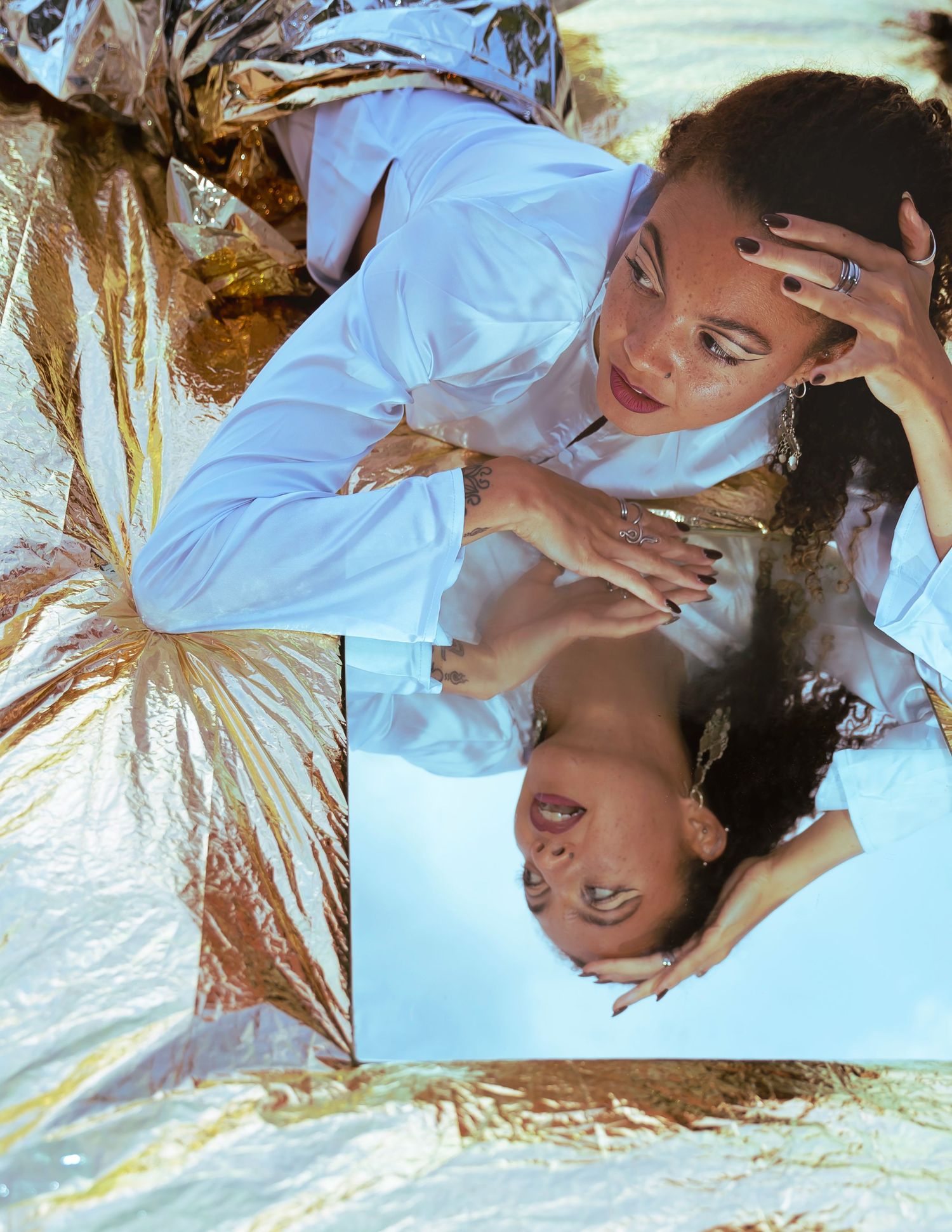 Woman in white shirt lying beside a mirror reflection on a gold and silver metallic surface.