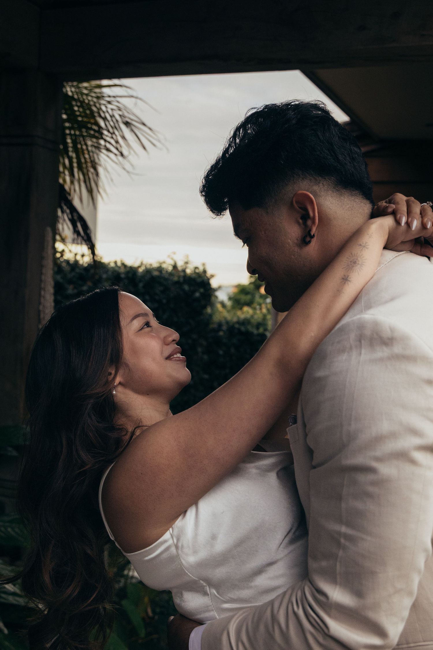 A couple in white embraces romantically outdoors, the woman smiling up at the man surrounded by lush greenery.