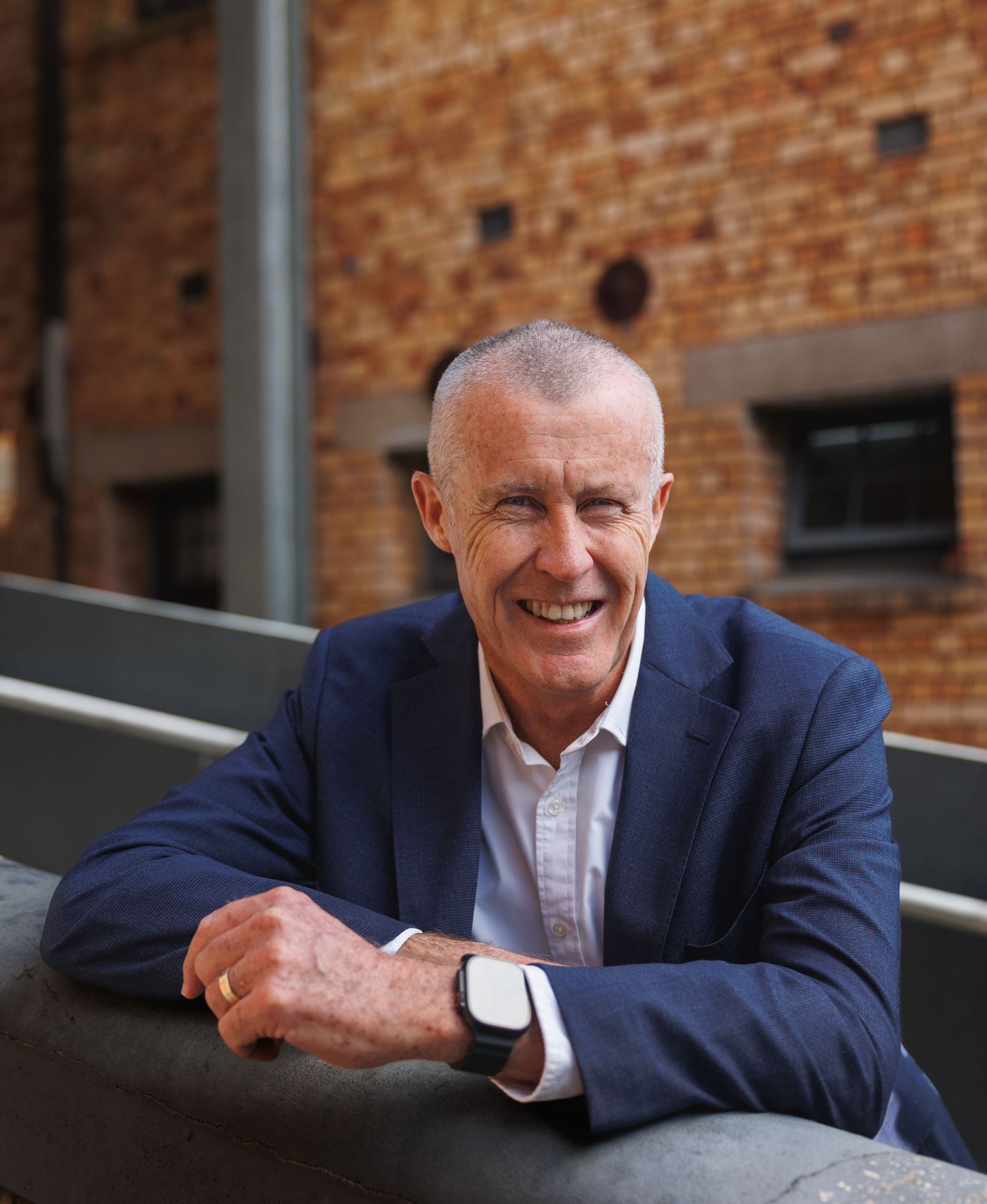 Smiling middle-aged man in a blue suit leaning on a railing outdoors with brick building background.