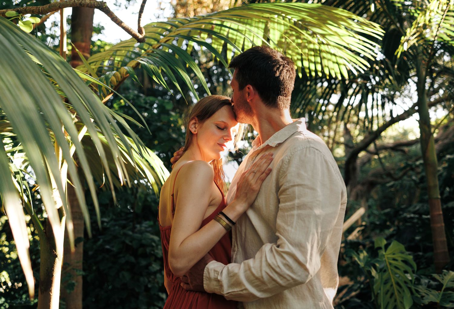 Romantic couple embracing closely among lush tropical palm trees in golden light.