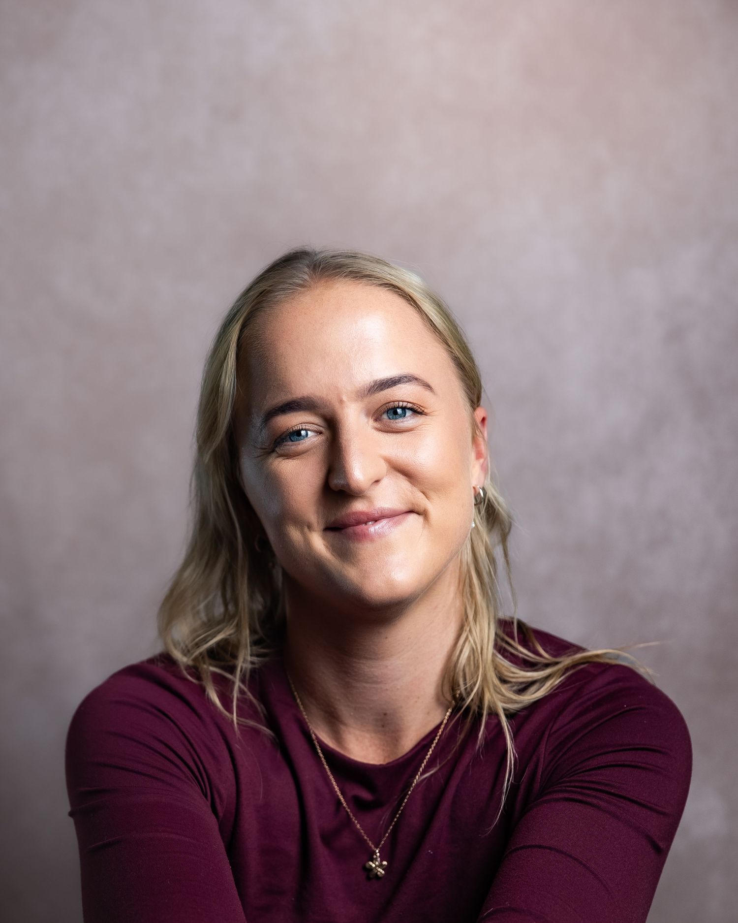 Smiling blonde woman in maroon top with gold necklace against neutral studio background.