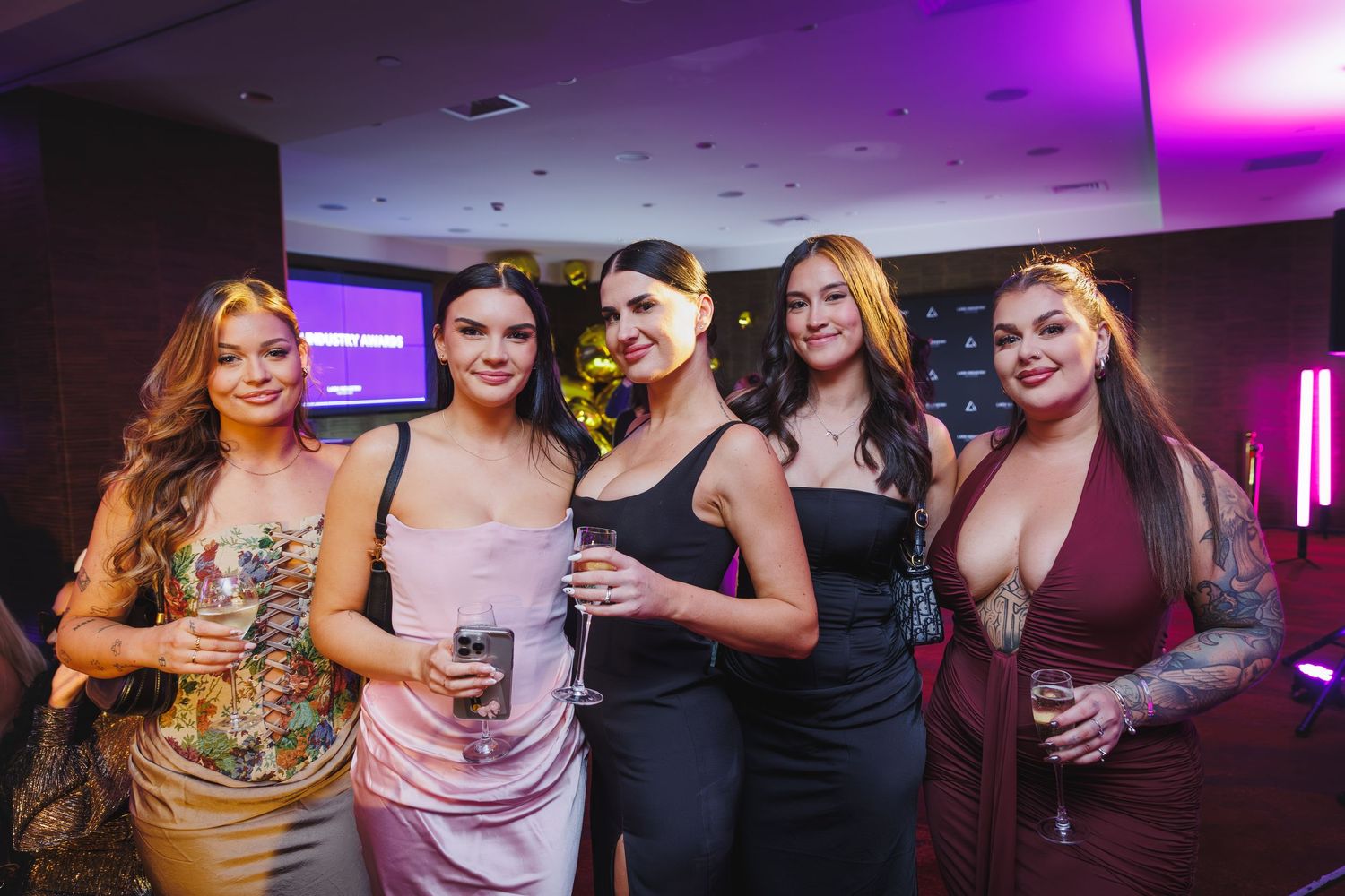 Five stylish women smiling together at an upscale evening event with purple lighting backdrop.