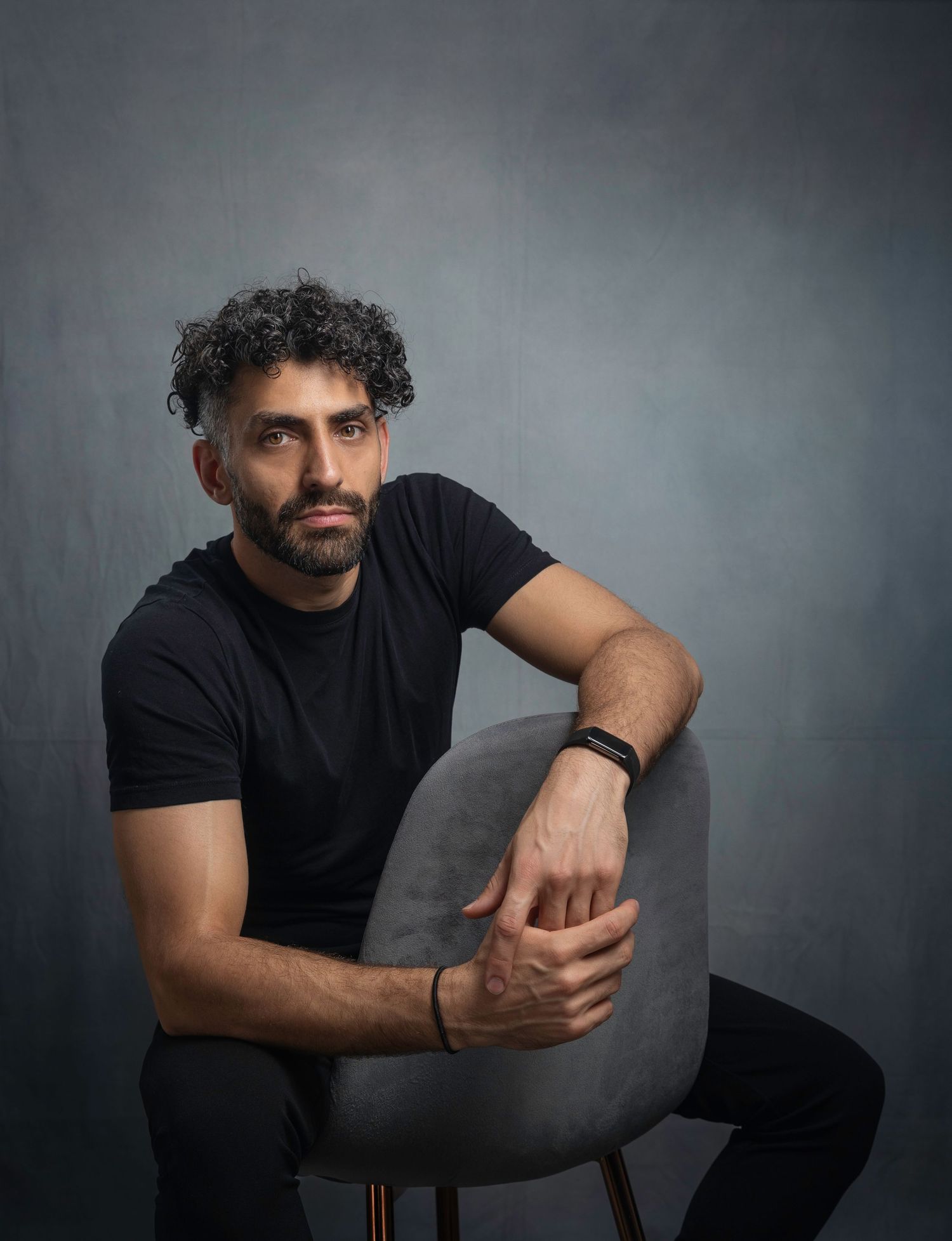 Bearded man with curly hair sitting on chair in black t-shirt against gray backdrop.