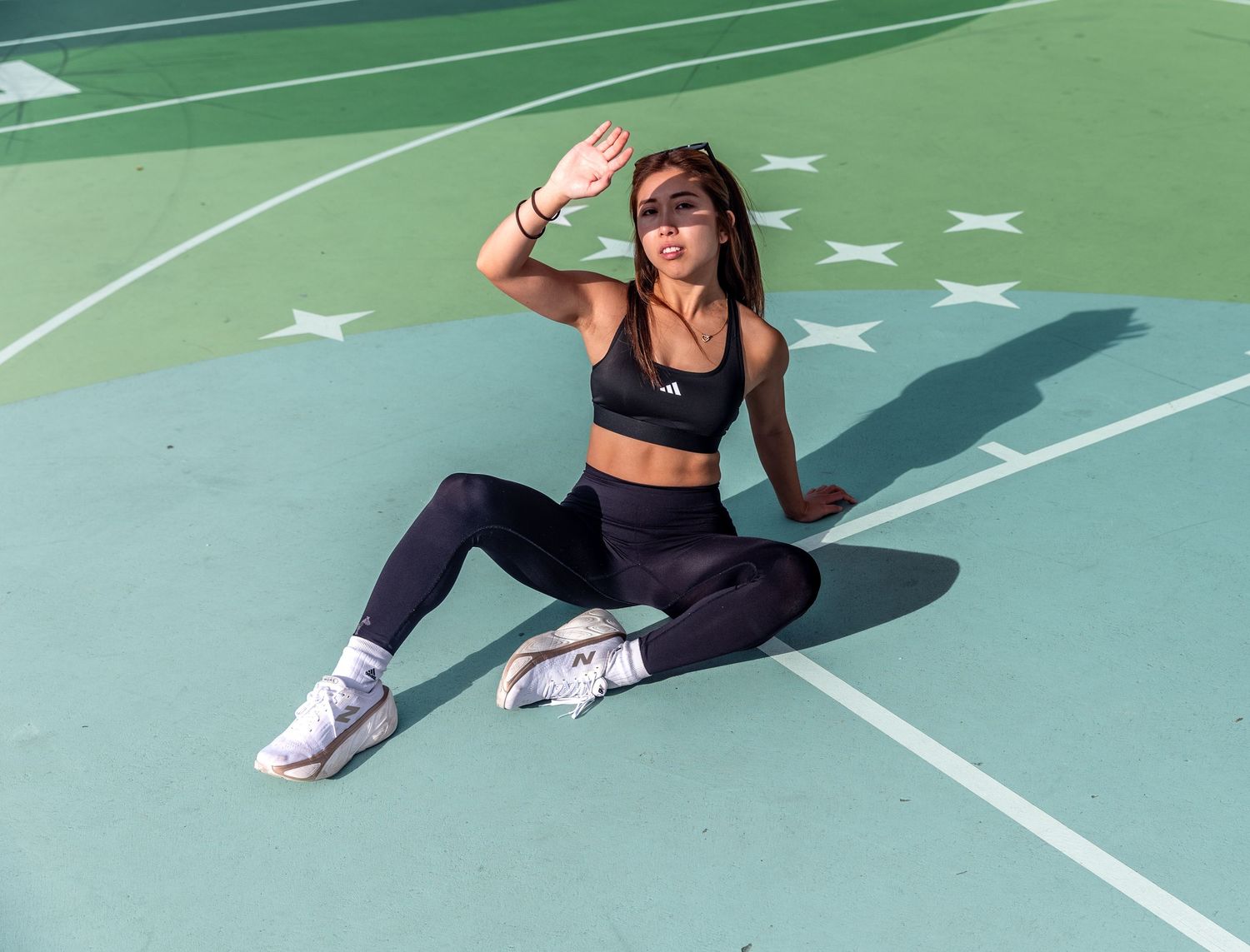 Athletic woman in black activewear sitting on a green sports court shielding her eyes from the sun.