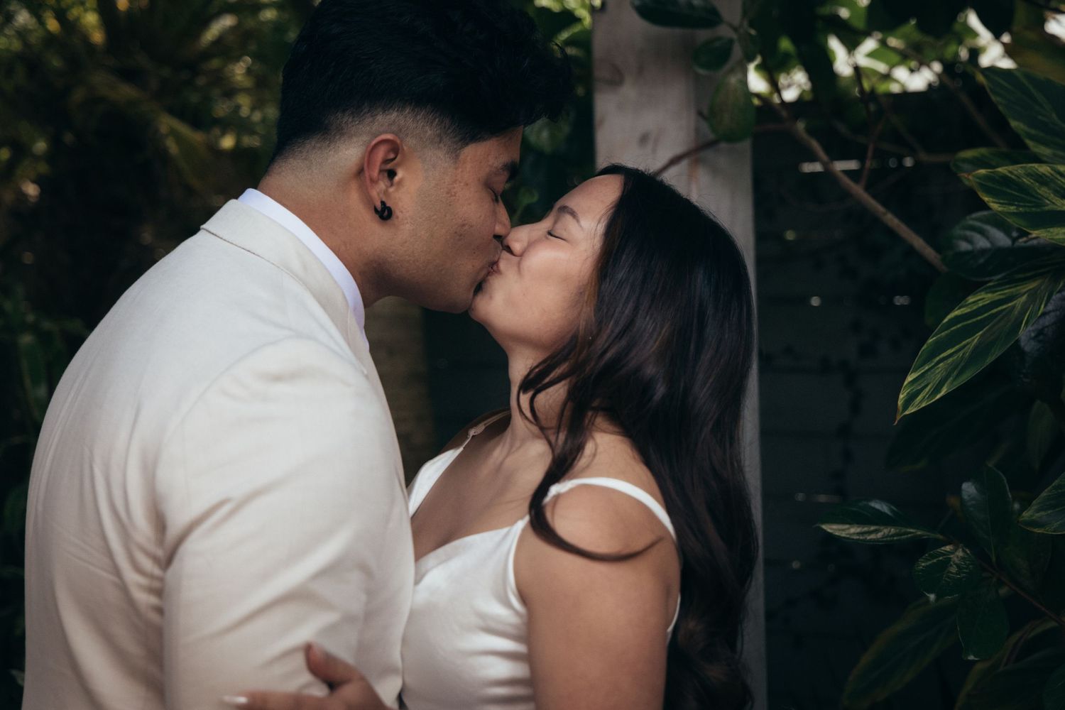 Couple sharing a romantic kiss outdoors surrounded by lush tropical greenery at their wedding.