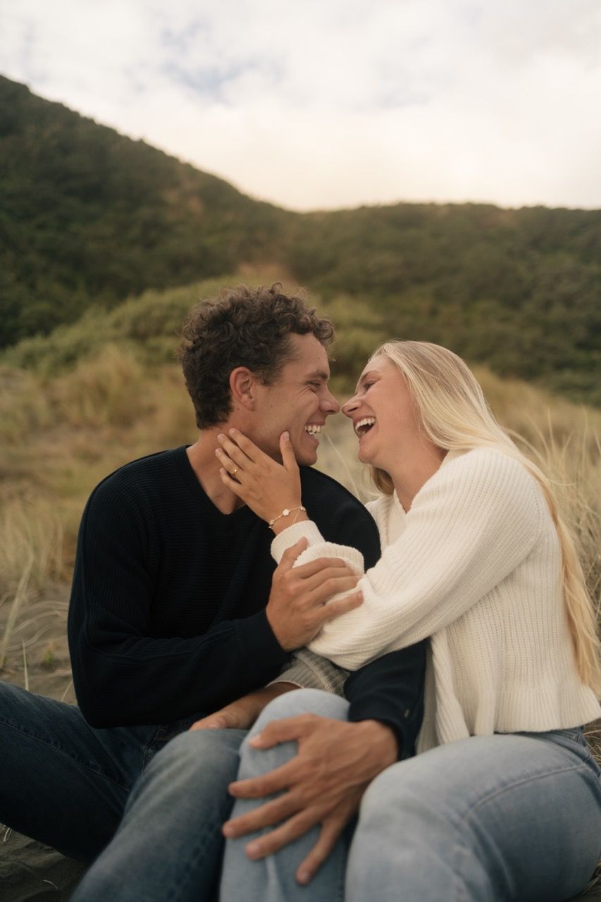 A laughing couple sits outdoors near green hills, sharing a joyful moment in casual sweaters and jeans.