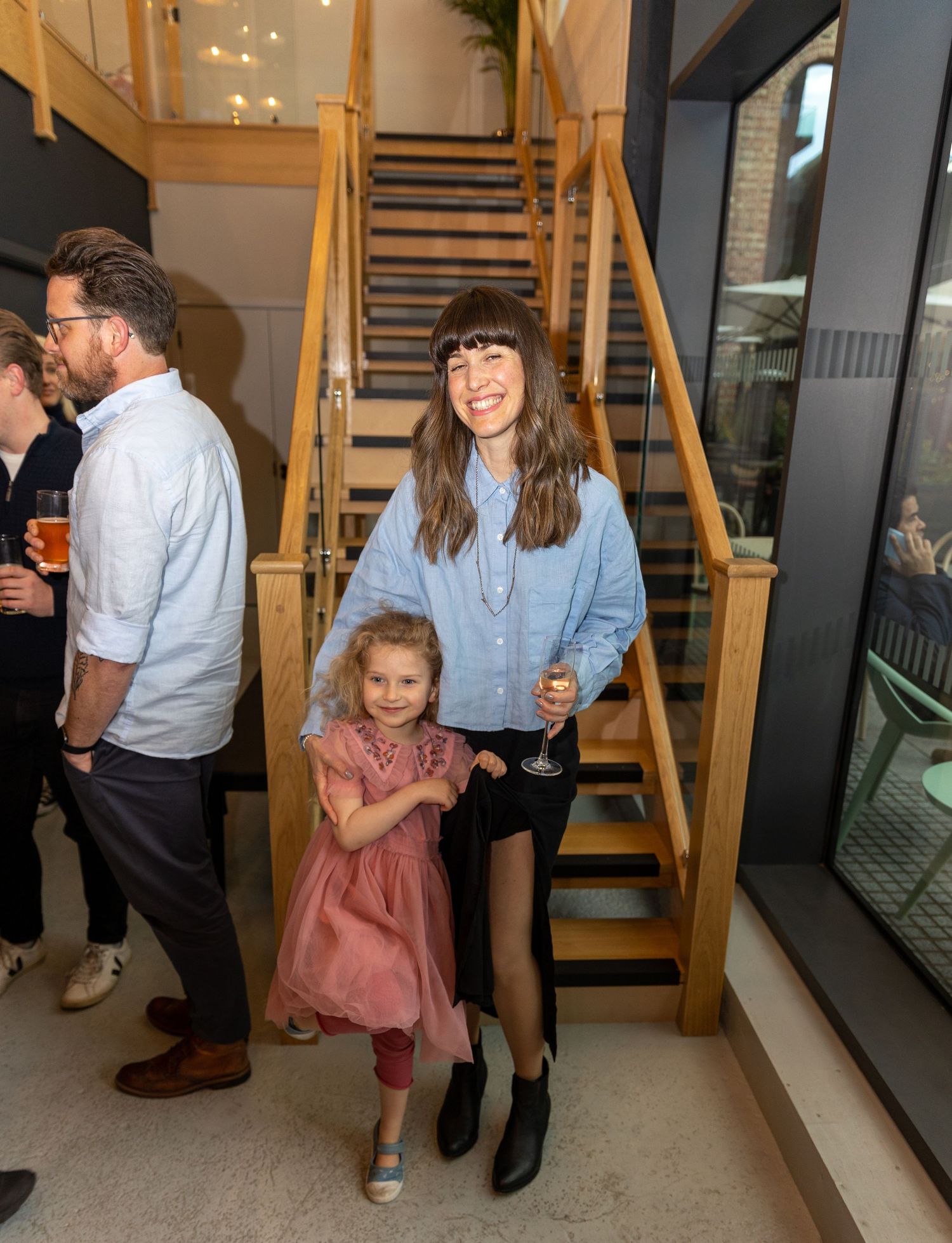 Woman and young girl in pink dress smiling near wooden staircase at an indoor social event.