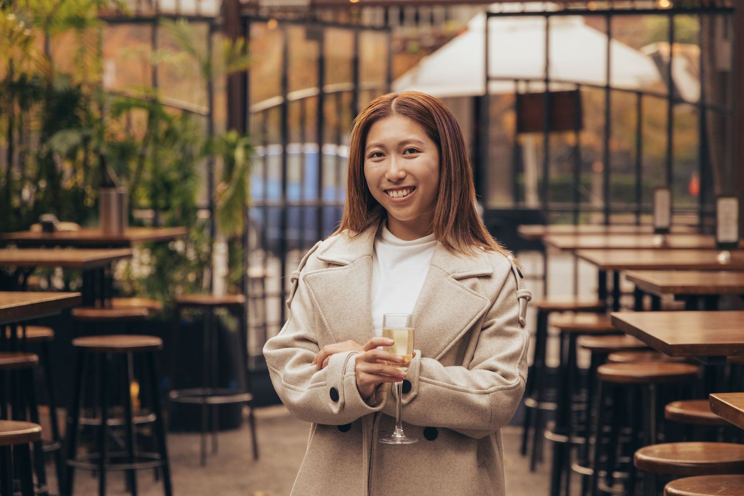 Happy Asian woman in beige coat holding white wine in an outdoor bar setting.