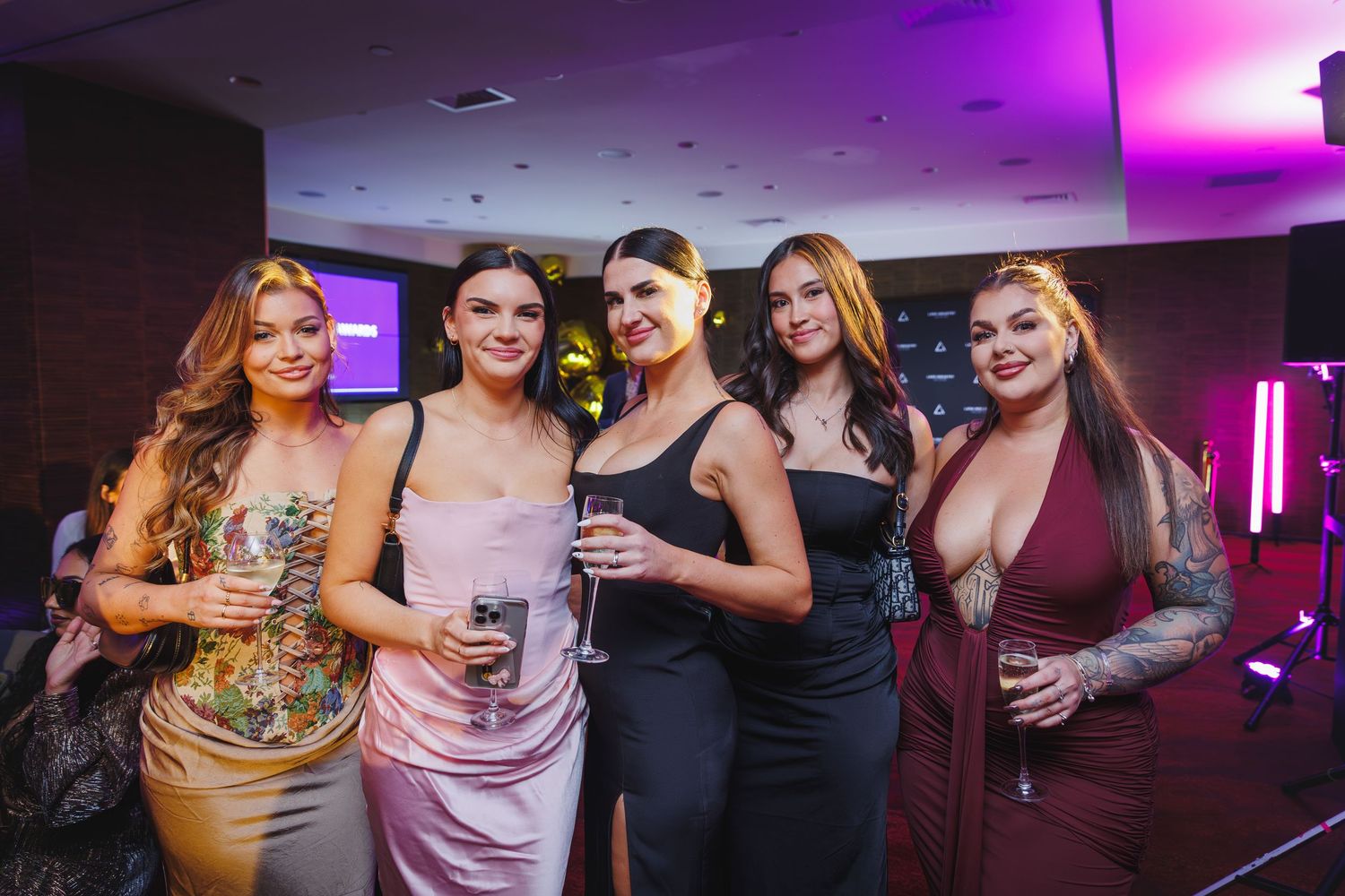 Five glamorous women posing together at a stylish evening event with purple ambient lighting.
