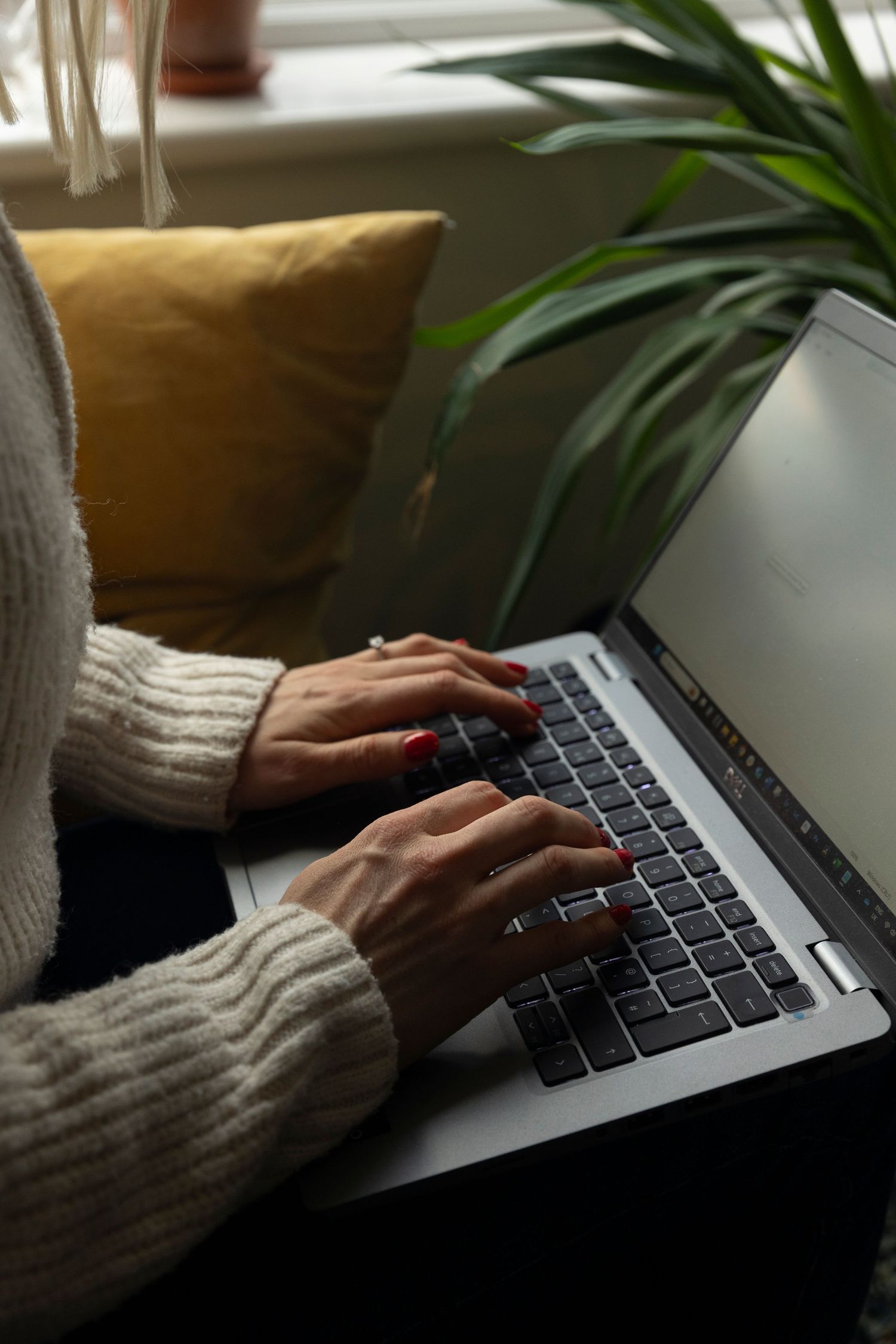 Close-up of hands with red nails typing on laptop keyboard near window with yellow cushion.