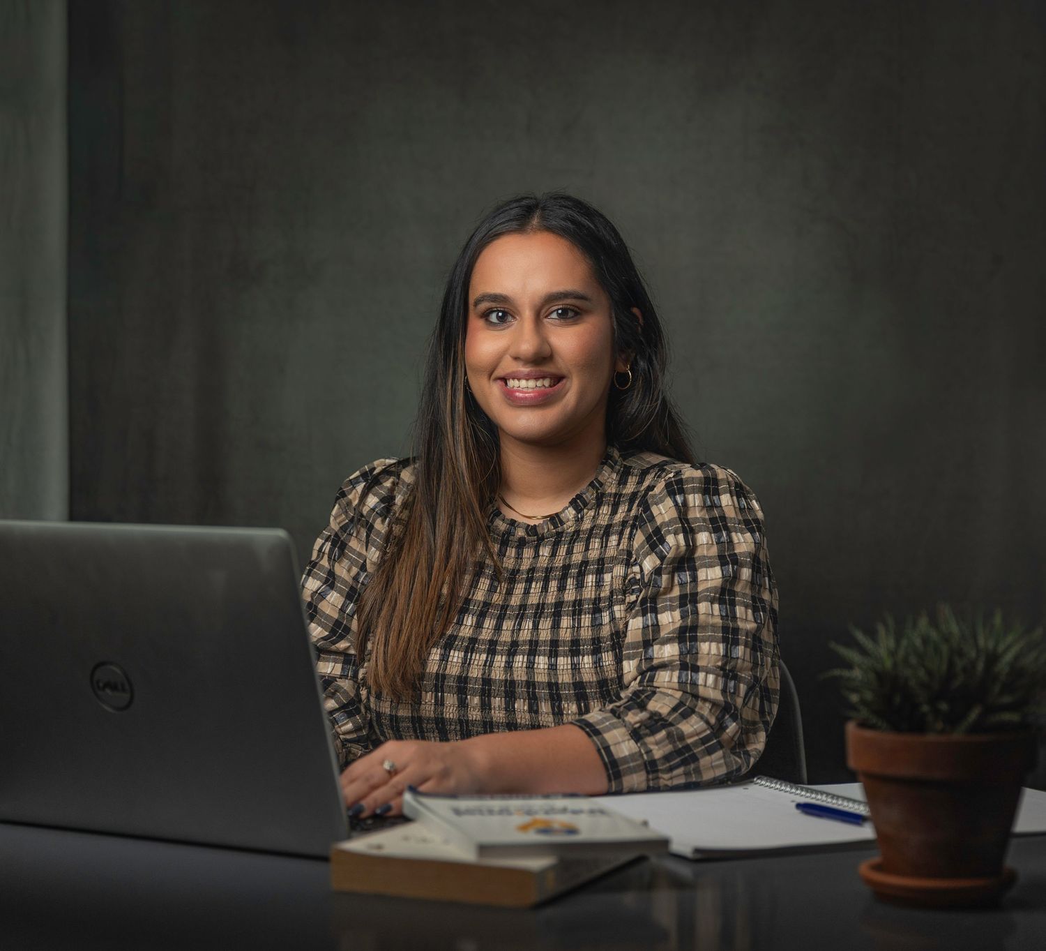 Young woman smiling at desk with laptop, notebook and plant in a professional office setting.