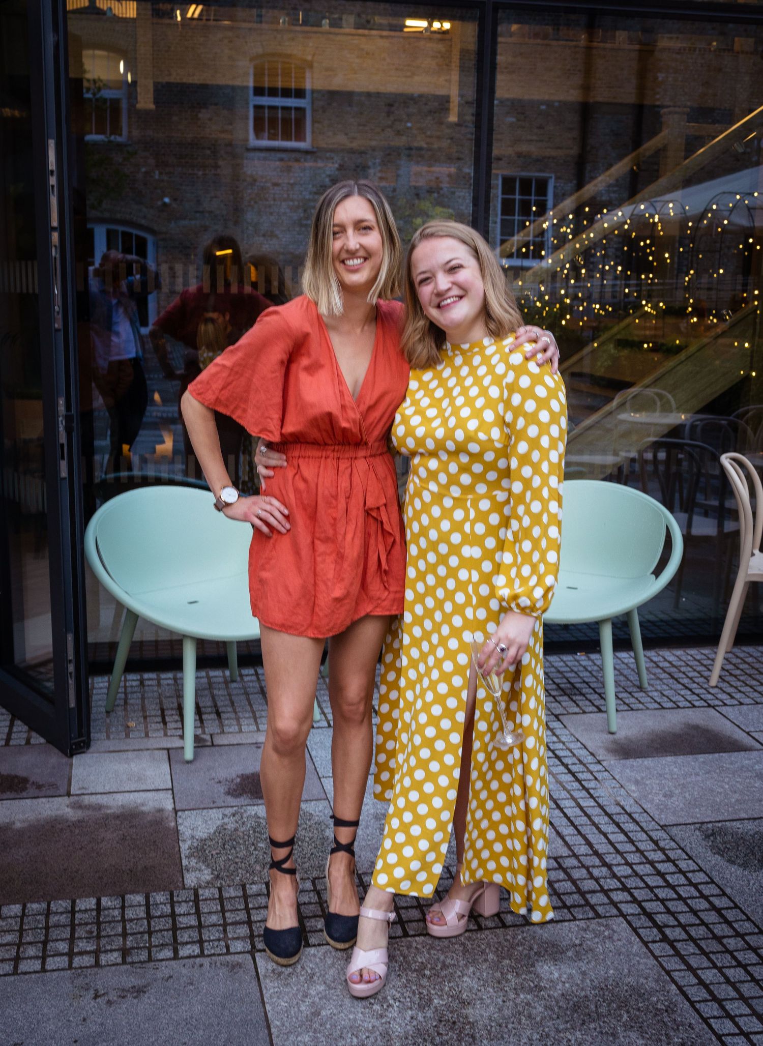 Two smiling women in colorful summer dresses posing together outside a modern restaurant entrance.