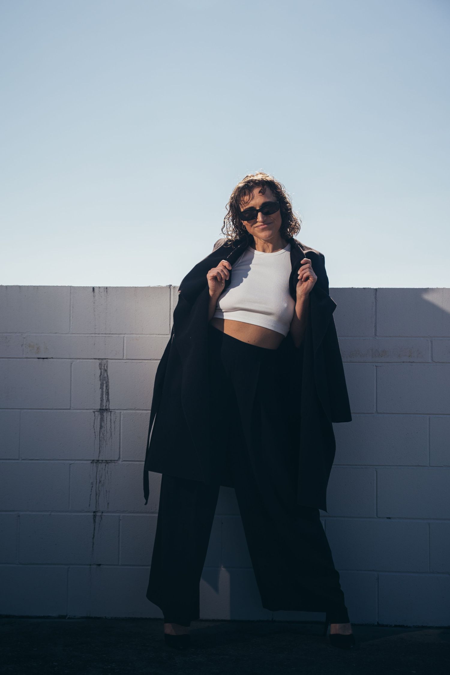Stylish woman in white crop top, black wide-leg pants and oversized coat poses against white brick wall.