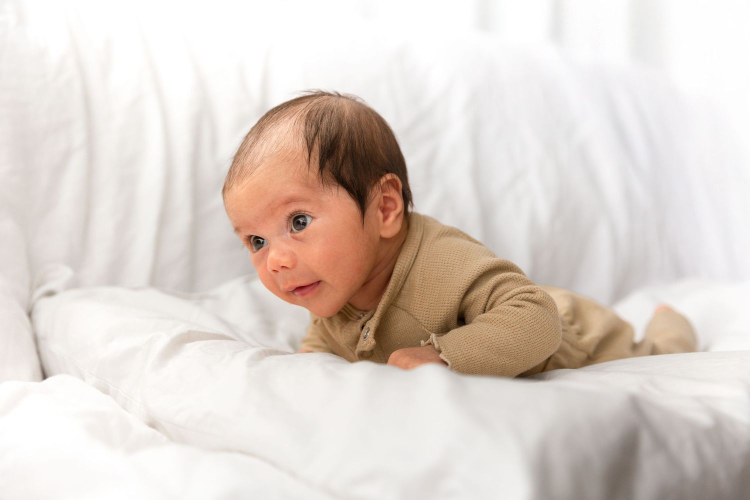 A curious newborn baby in a olive green onesie doing tummy time on a soft white blanket.