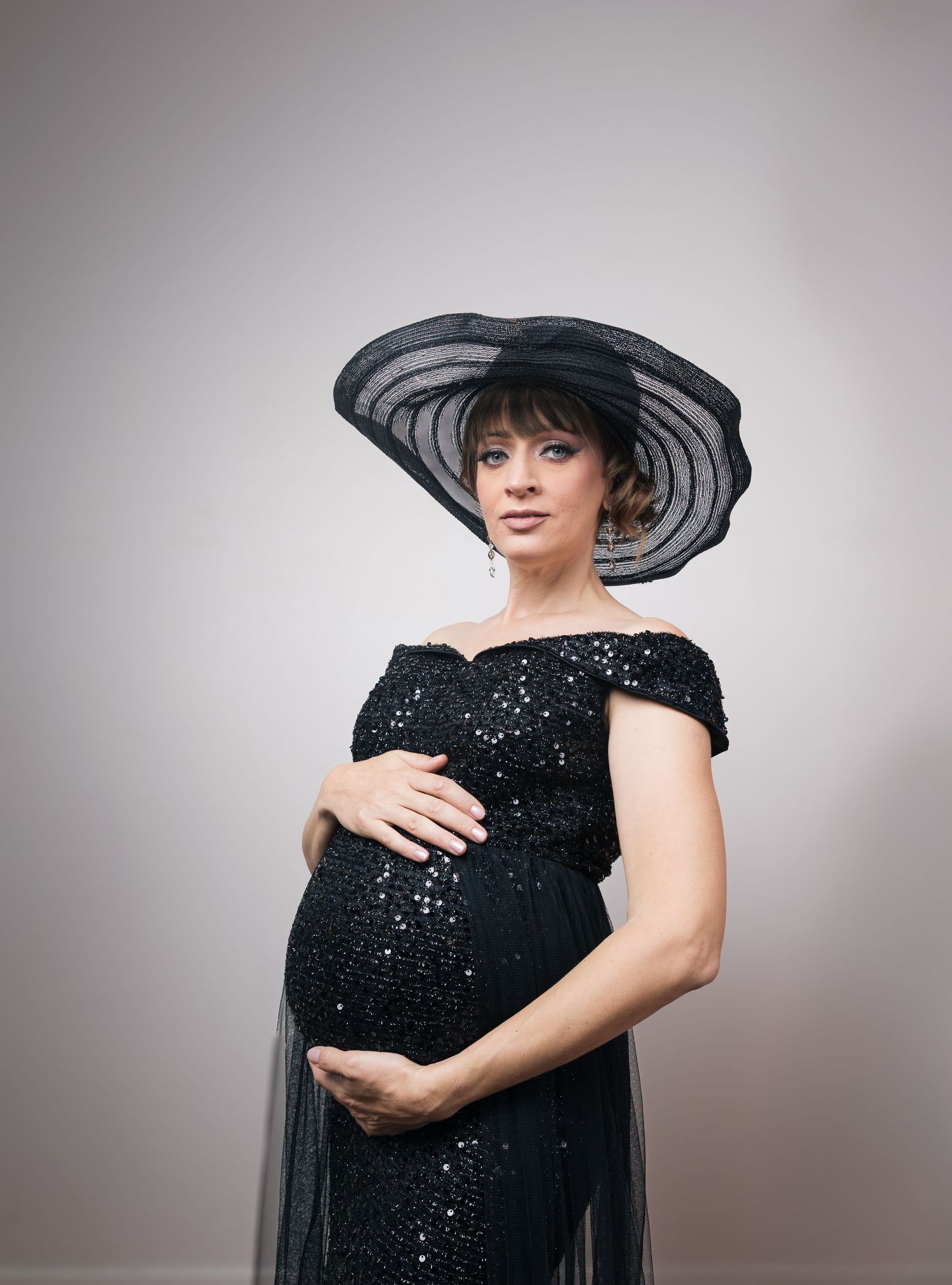 Pregnant woman in black sequin off-shoulder gown and wide-brim hat cradling baby bump in elegant maternity portrait.