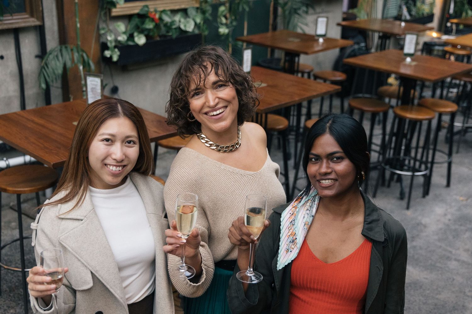 Three diverse women smiling and holding champagne glasses at an outdoor restaurant patio.