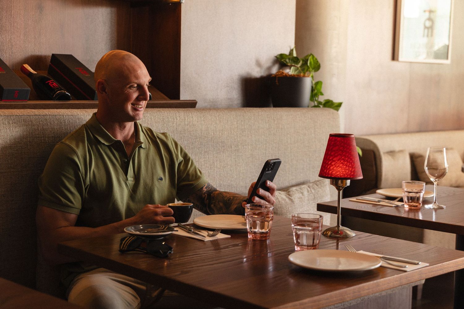 Bald man smiling at his phone while dining alone at a restaurant table with a red lamp.