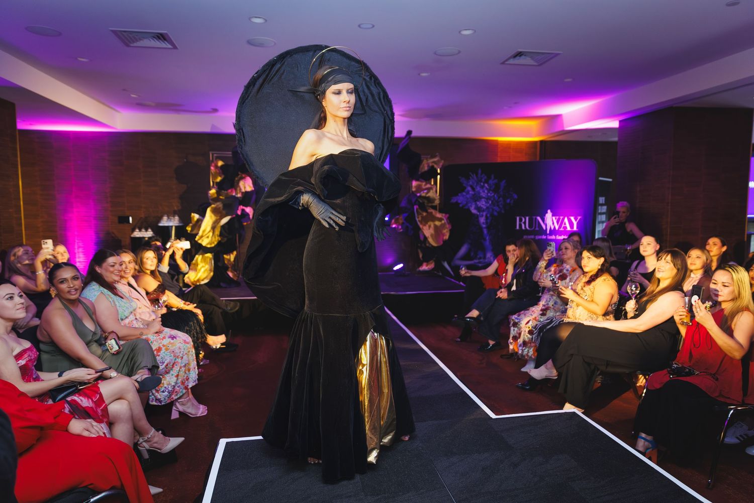 Model in a dramatic black gown with large hat walks the runway at the Lash Industry Awards show.