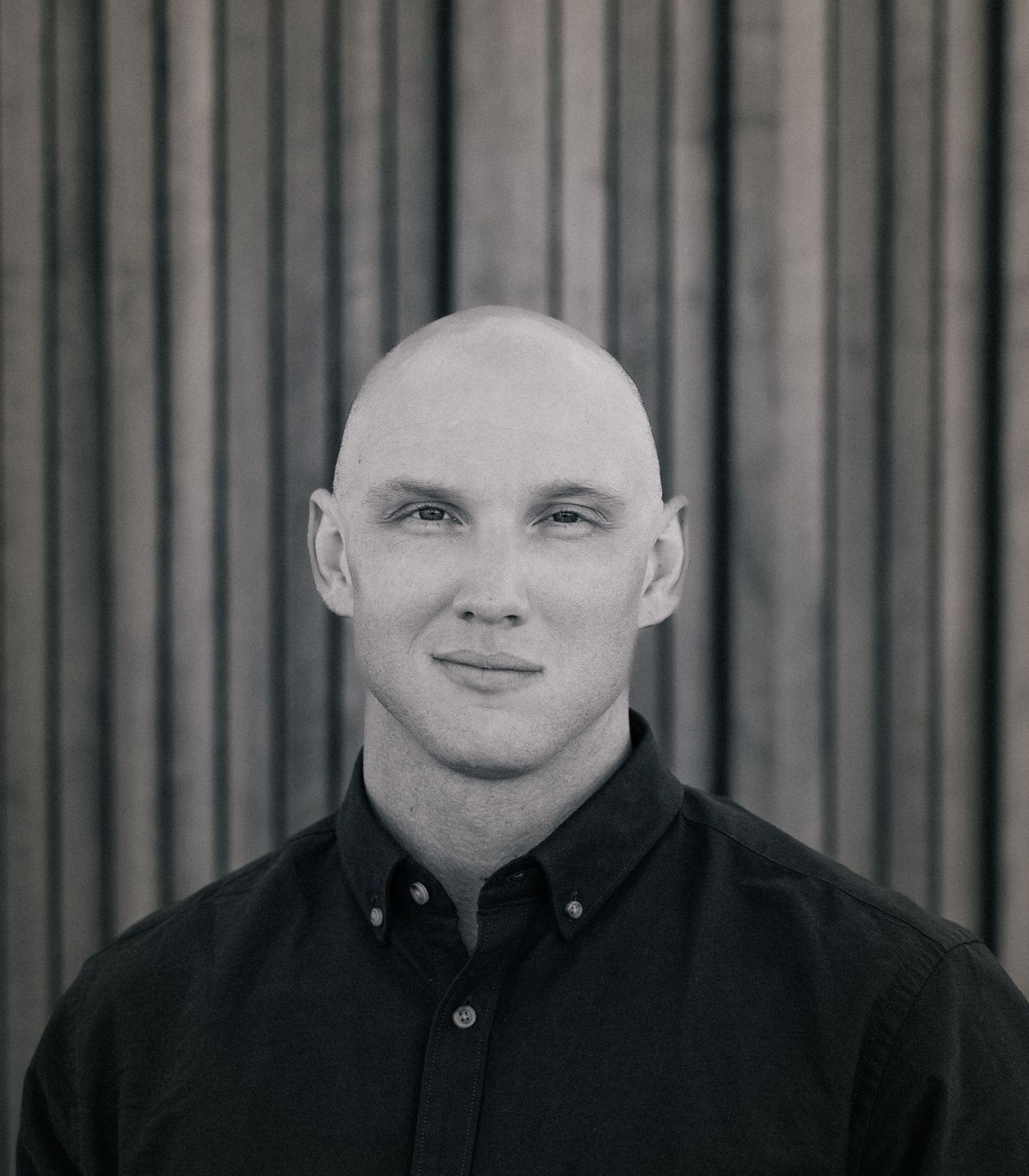 Black and white portrait of a bald man in a dark shirt smiling against a striped background.