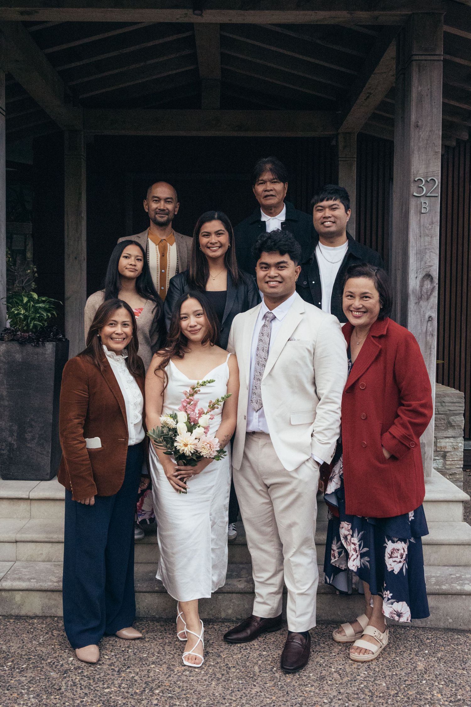Wedding couple poses with nine family members outside a venue entrance surrounded by tropical foliage.