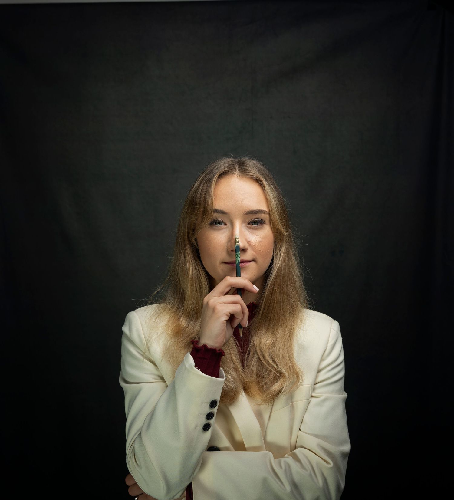 Blonde woman in white blazer holding a pen thoughtfully against a dark studio background.